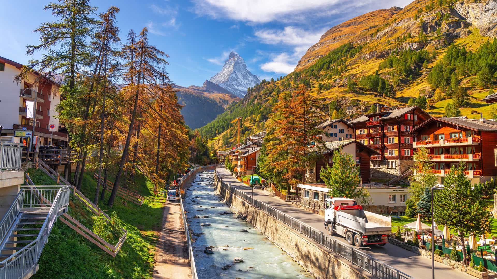 A vibrant autumn view of the Zermatt village in Switzerland, with the iconic Matterhorn mountain in the background, surrounded by colorful trees and a flowing river.