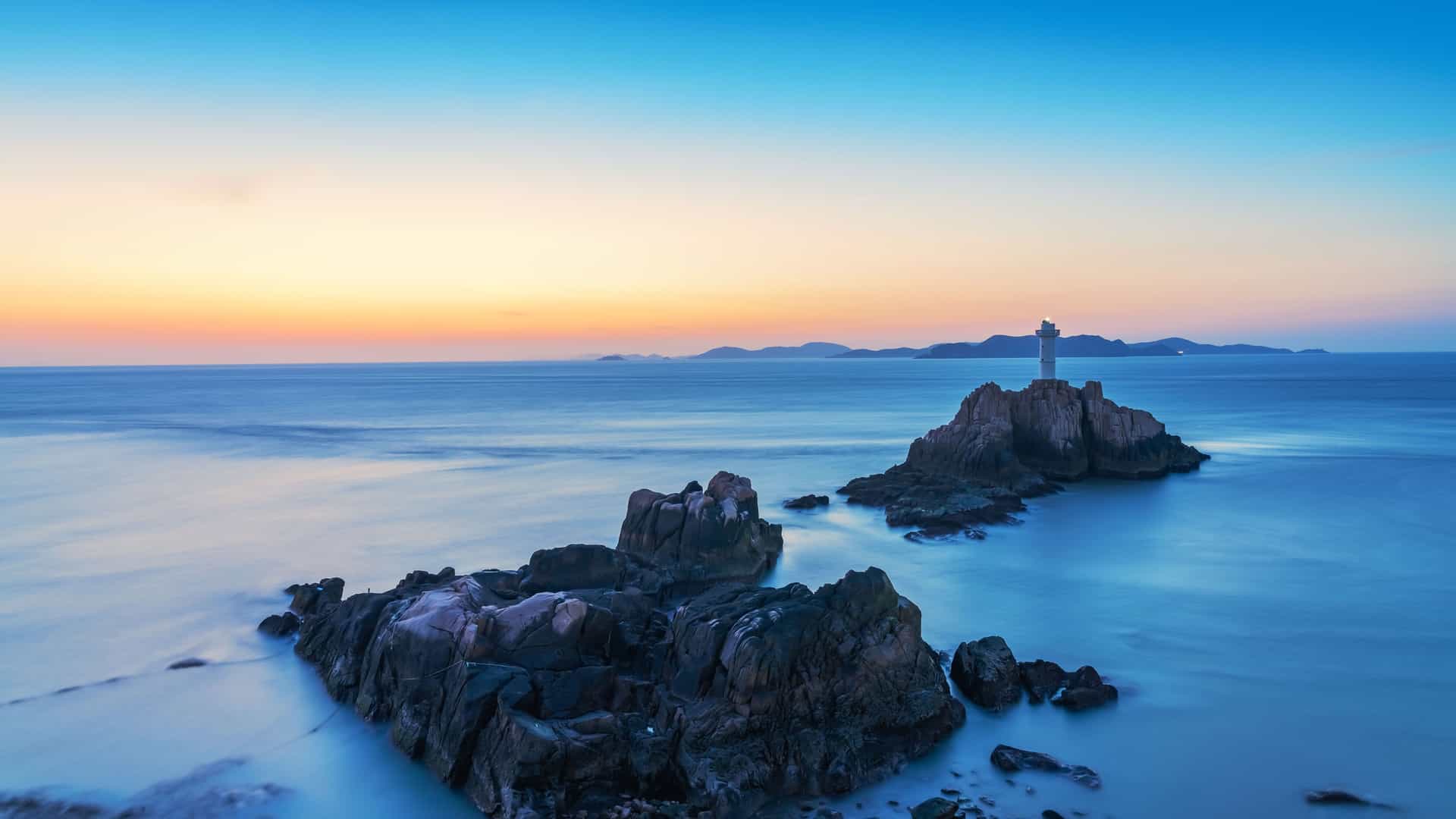 A breathtaking dusk shot of a small lighthouse on a rocky outcrop in the sea off the coast of Zhoushan, China, with a smooth water effect and a clear sky.