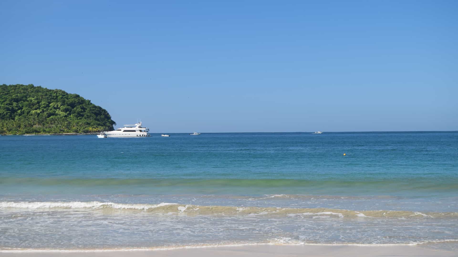 A tranquil beach scene in Zihuatanejo with gentle waves rolling onto sandy shore, turquoise water dotted with a white yacht and smaller boats, and a lush green island extending into the sea beneath a clear blue sky.