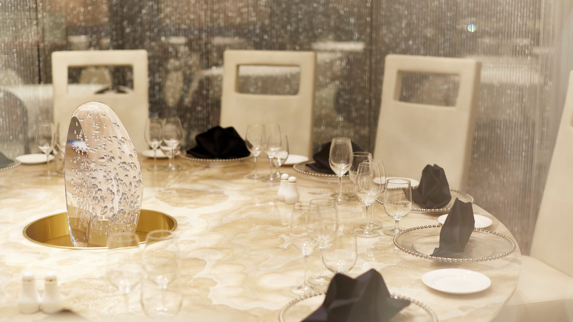 A close-up view of an elegant round dining table set with pristine place settings, black napkins, and a glowing bubble-glass centerpiece, surrounded by shimmering silver beaded curtains.