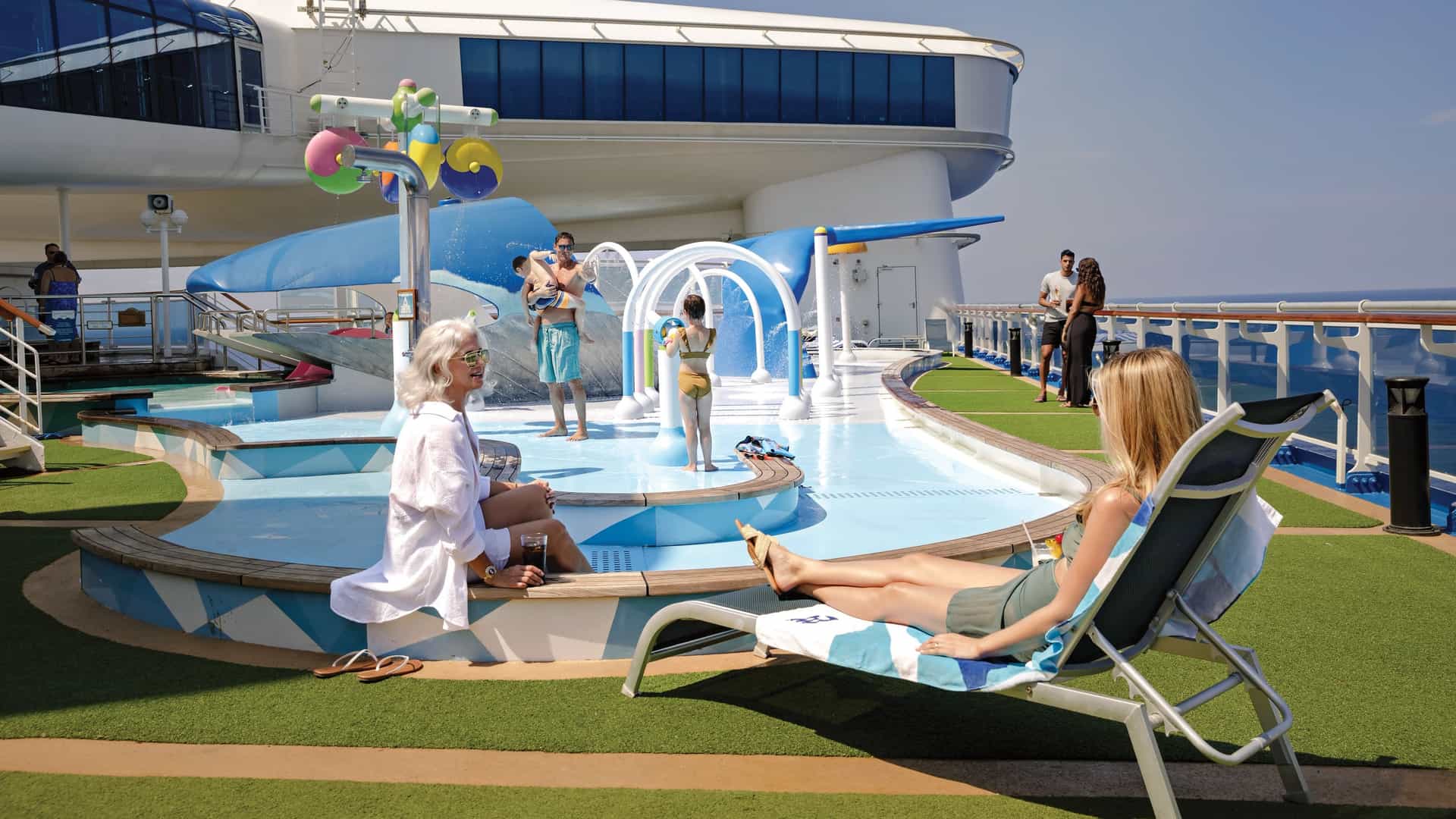 A family enjoying an outdoor kids' water park and splash pad area on a cruise ship deck, with adults lounging in the foreground and children playing under water features against an ocean backdrop.