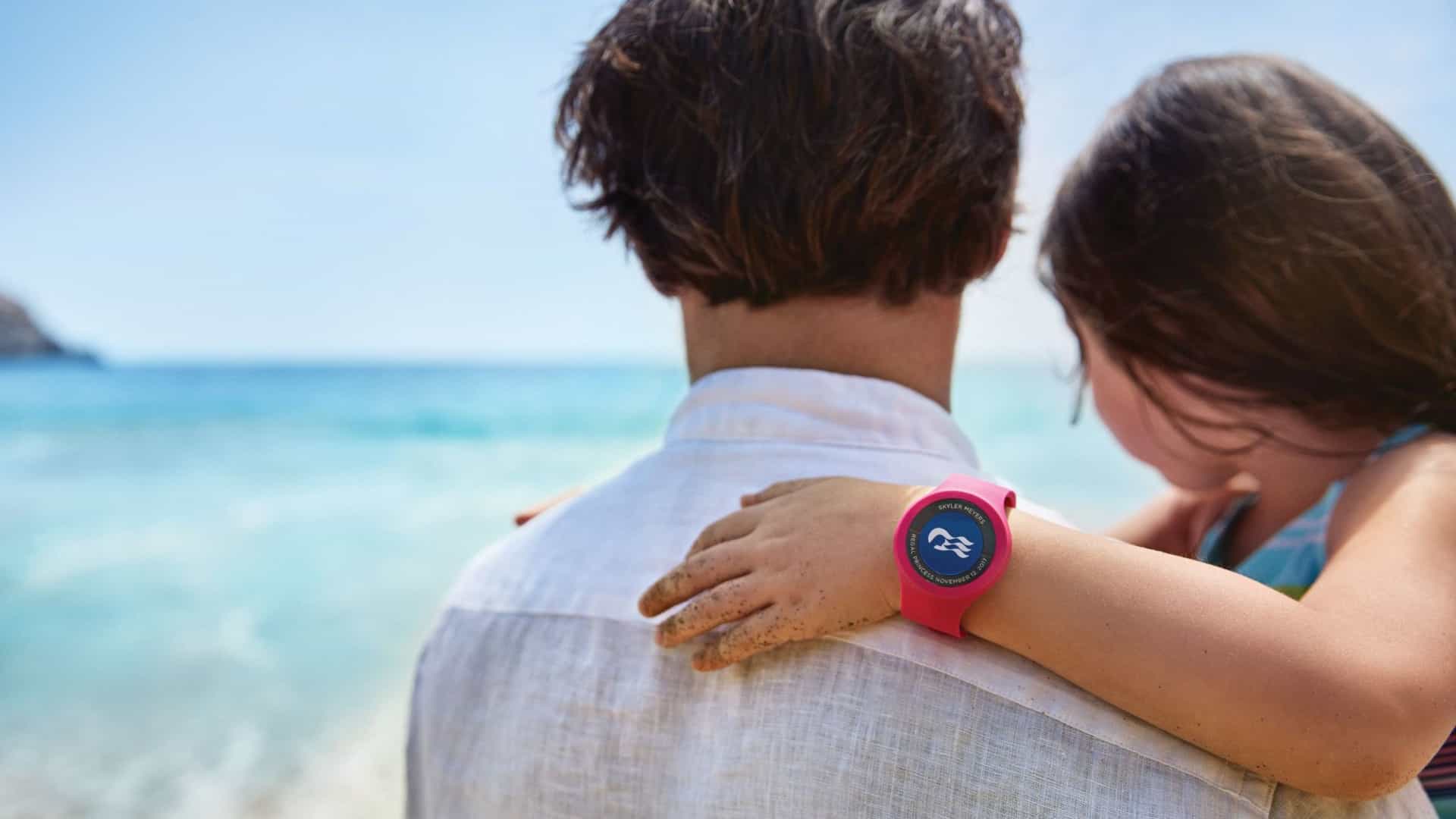 A father holds his child wearing a pink Princess Cruises Medallion wristband while looking out at the ocean during a beach vacation.