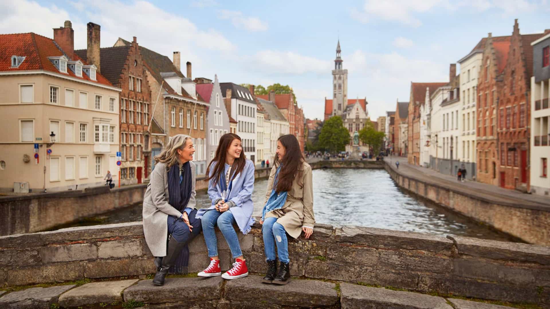 Three women sit on a charming stone bridge overlooking a historic canal in a Northern European city like Bruges, a picturesque stop on a Princess Cruises itinerary.