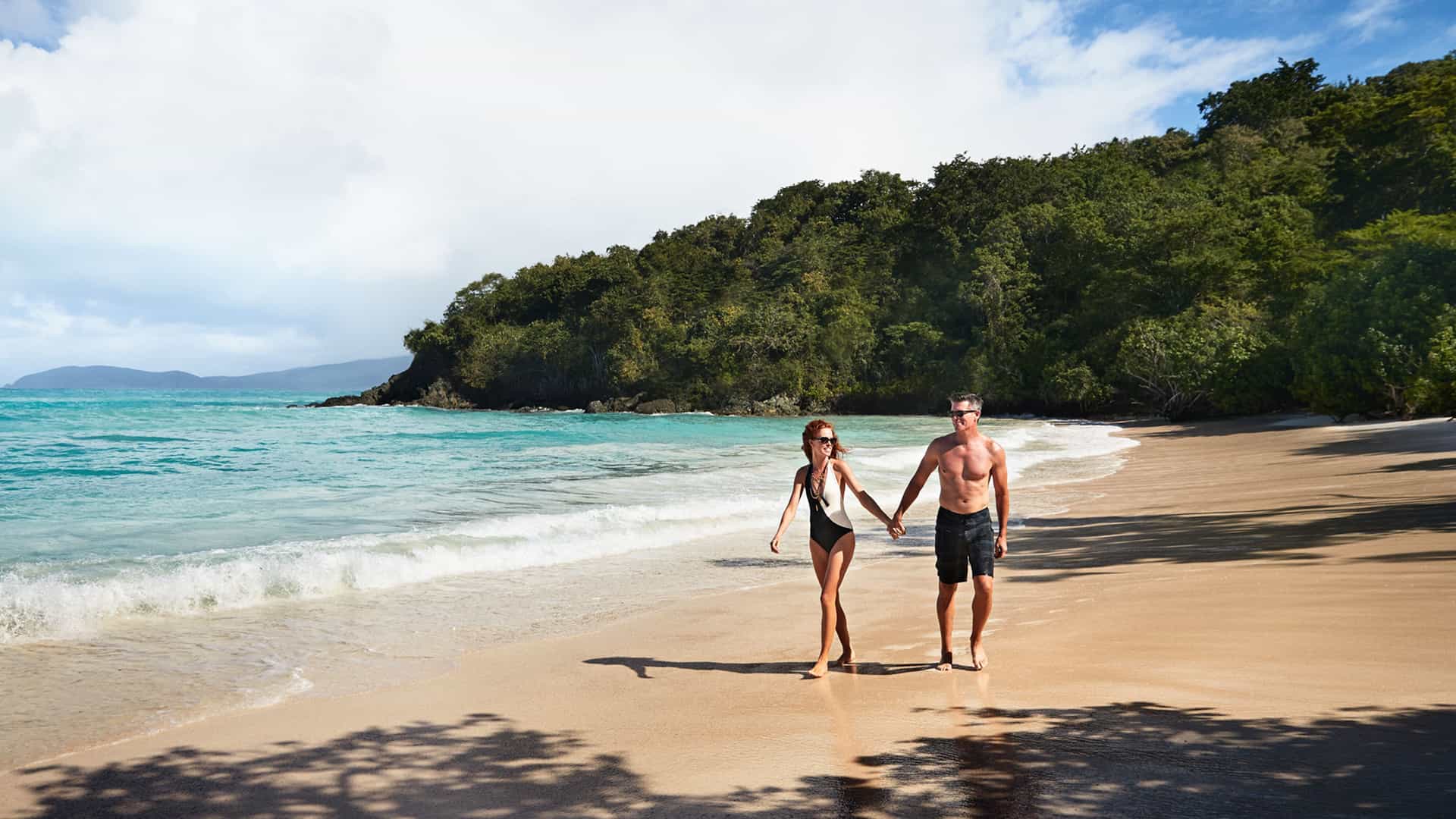 A couple in swimsuits walks hand-in-hand along a pristine sandy beach with turquoise water, a lush green hillside, and a partly cloudy sky, perfect for a Princess Cruises Southern Caribbean itinerary.