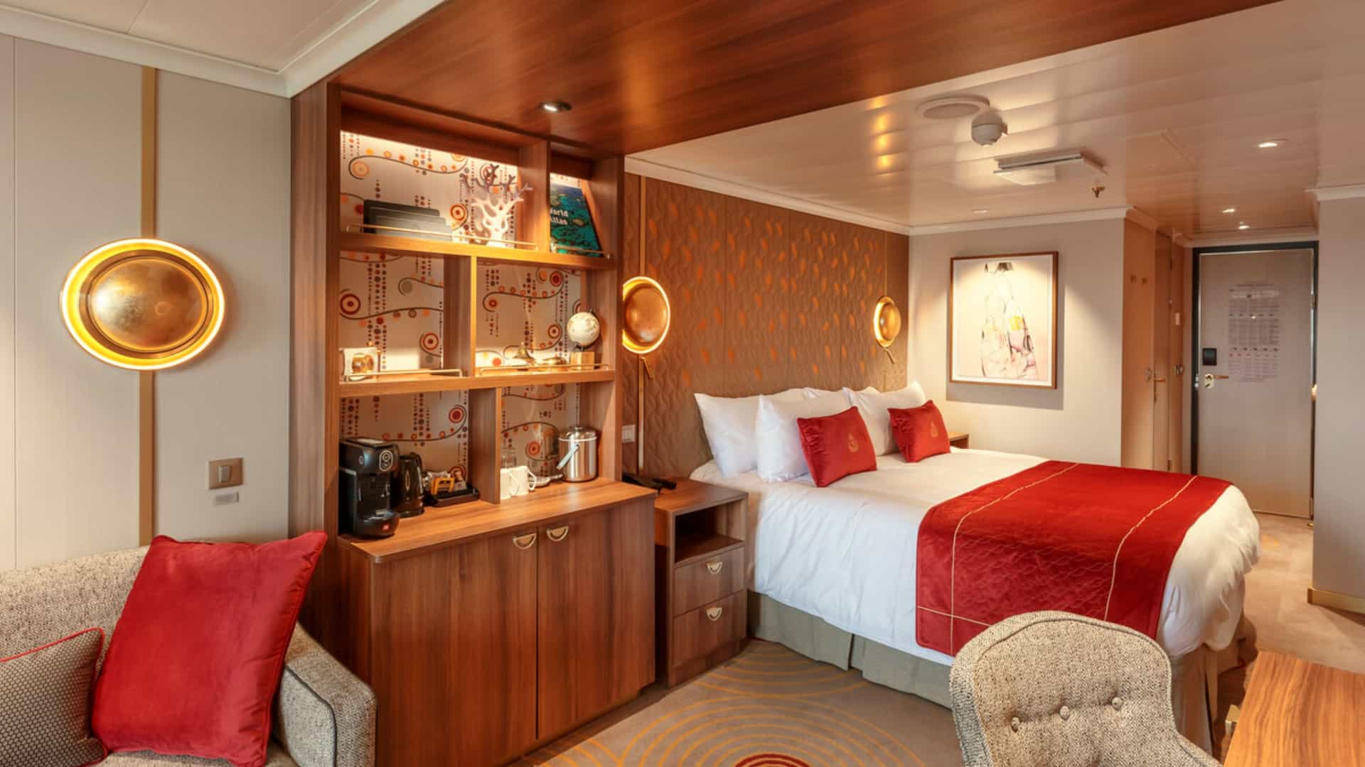 Elegant Princess Grill suite on Queen Anne showcasing a plush bed with rich red accents, custom wood cabinetry, and a fully stocked mini-bar setup.