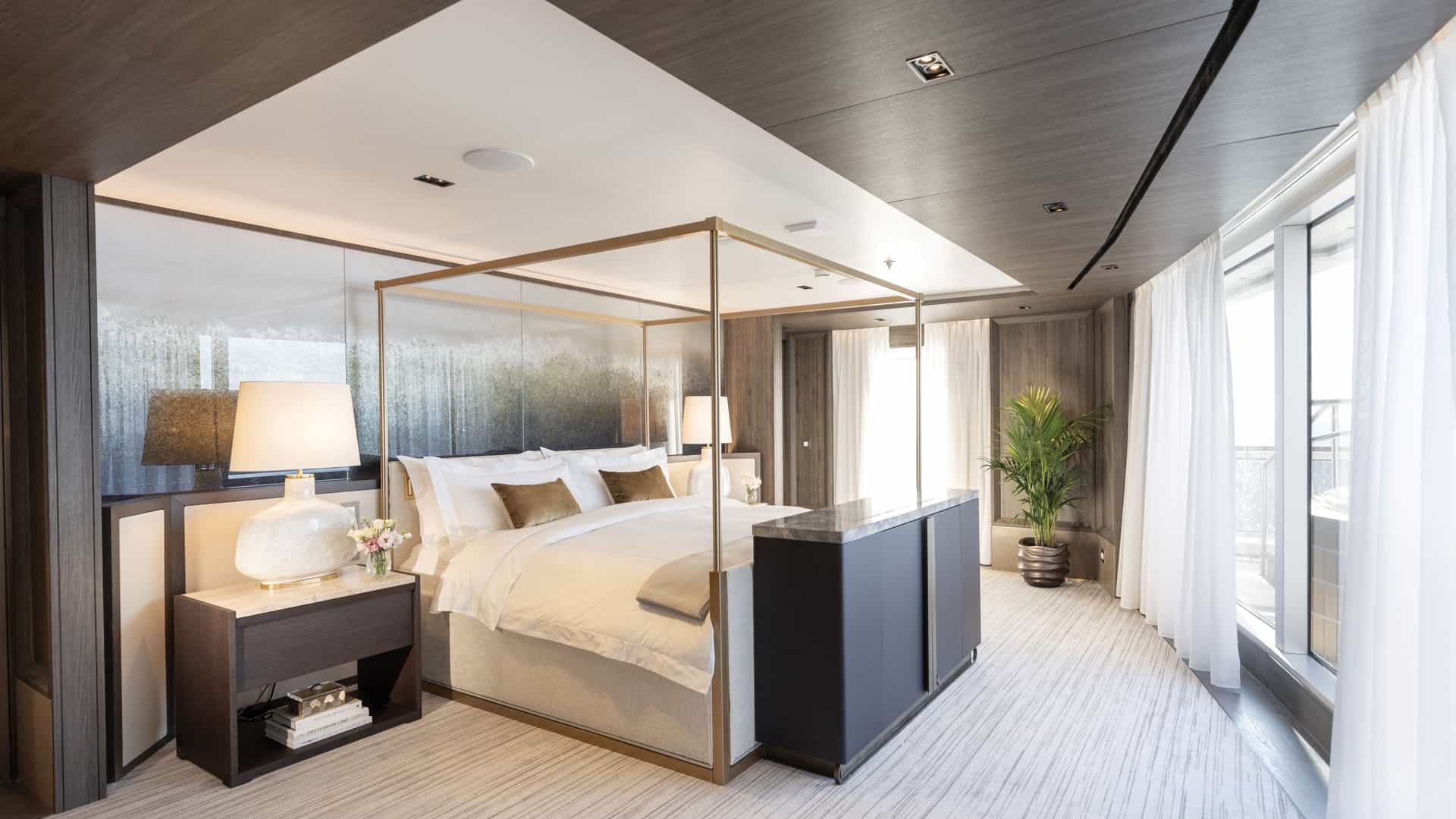 A wide shot showing a modern master bedroom suite aboard the Grandeur on Regent Seven Seas. Windows draped with linen and light shining through, showing the sharp angles of the bed and the decor of the room.