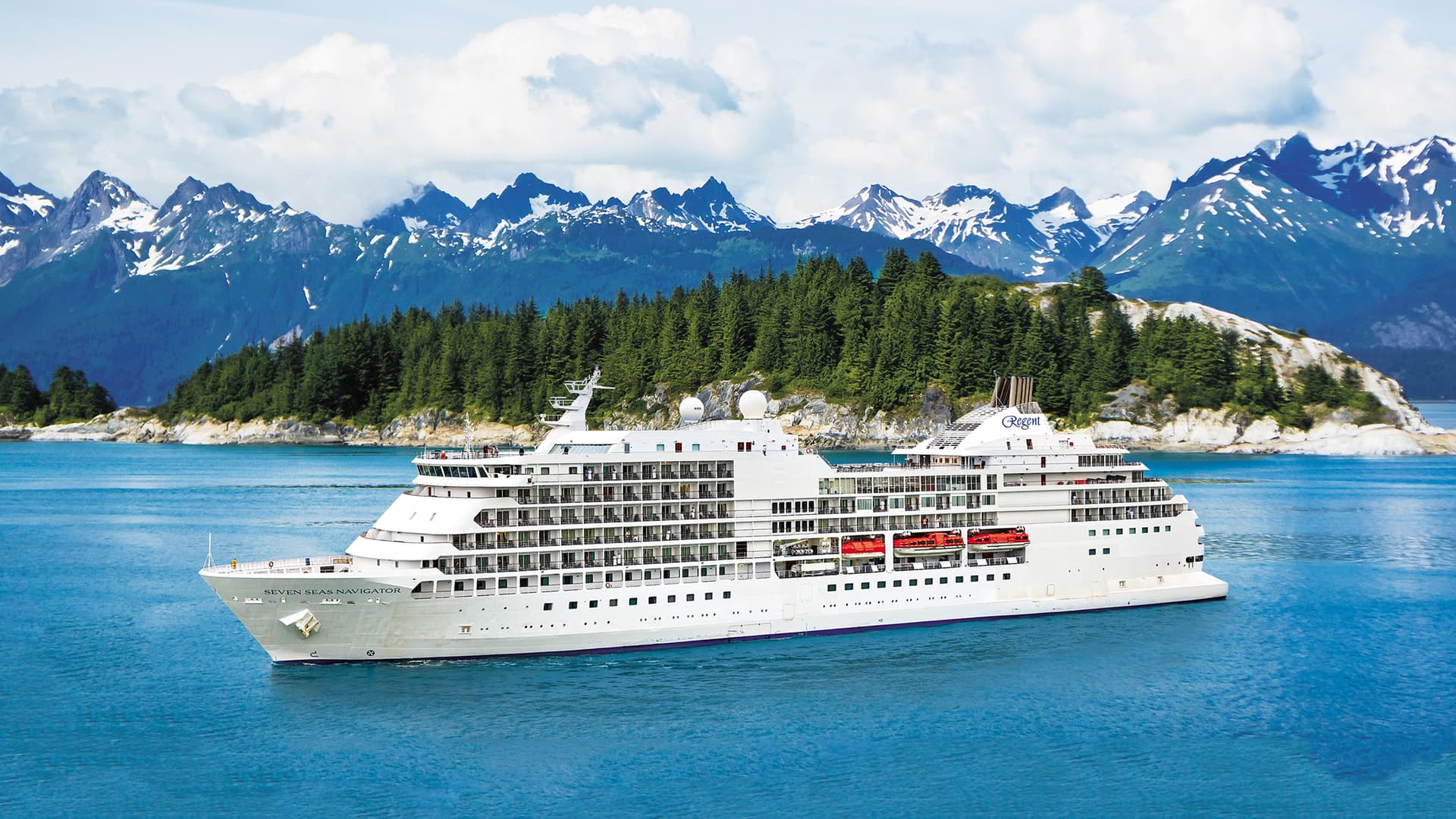 Regent Seven Seas Navigator Cruise Ship sailing in front of trees and snowy mountains.