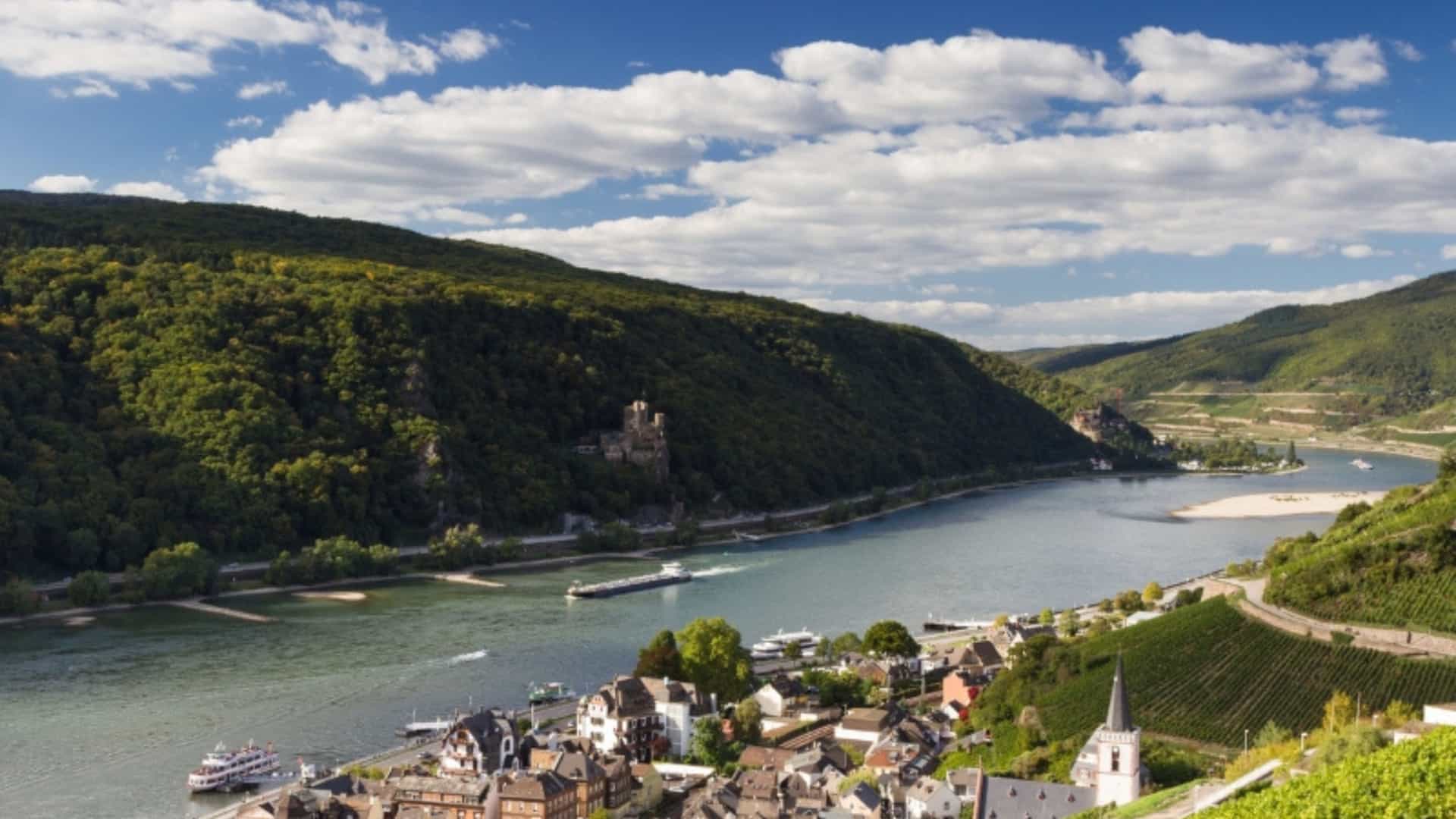 An elevated view of the Middle Rhine Valley in Germany, showcasing the Rhine River as it winds through a landscape of steep, green hills covered in vineyards and dense forests. A small village with a church steeple is nestled on the riverbank, and a barge is visible on the water. In the distance, a castle sits atop a forested hill, capturing the scenic beauty of the Rhine region.