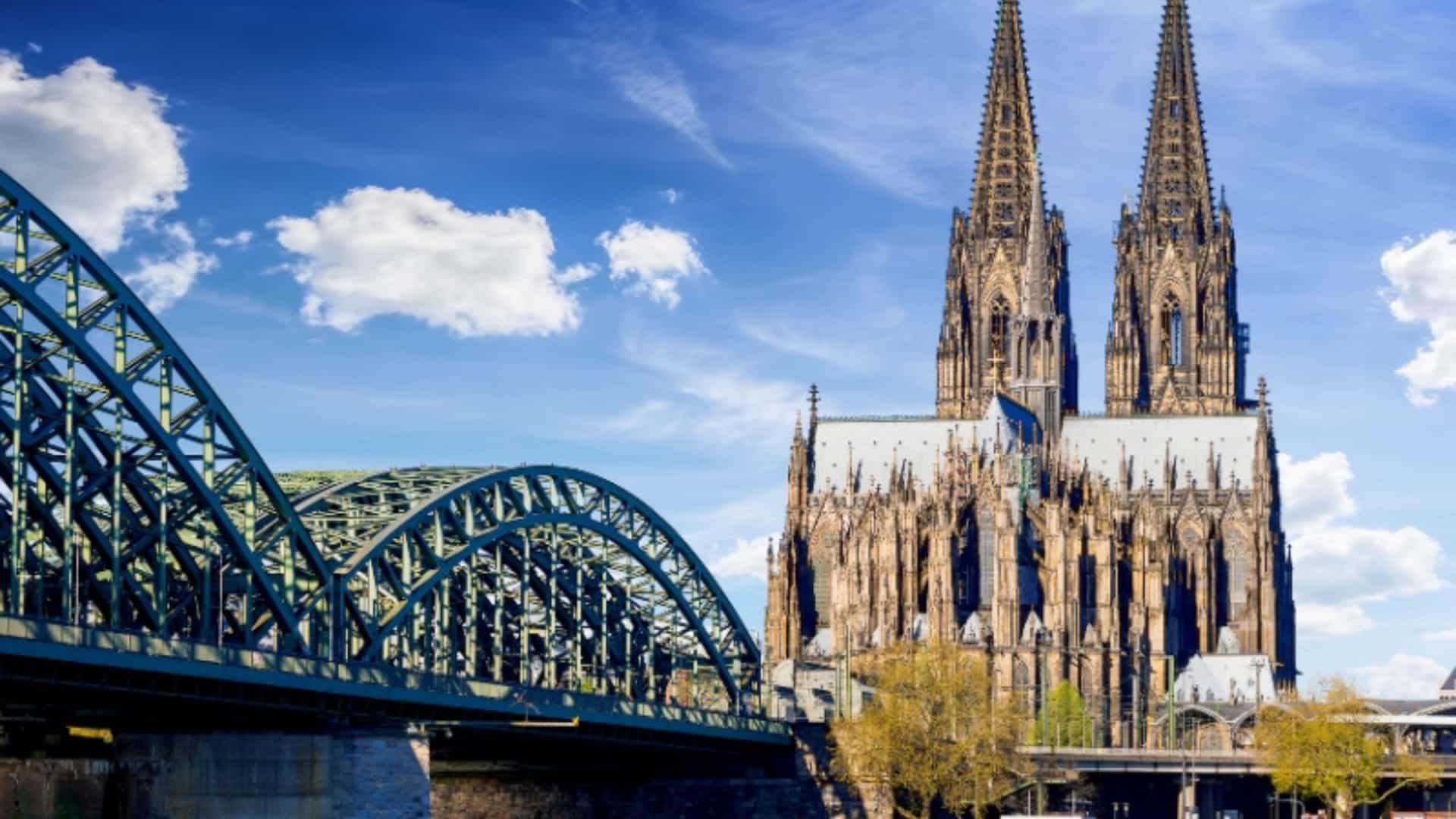 A view of the Cologne Cathedral and the Hohenzollern Bridge spanning the Rhine River in Germany. The gothic cathedral has two towering spires, and the bridge features a distinctive steel arch design. Small boats are docked on the river, and the sky is a bright blue with scattered clouds. The image captures the iconic architecture of a major city on the Rhine River.