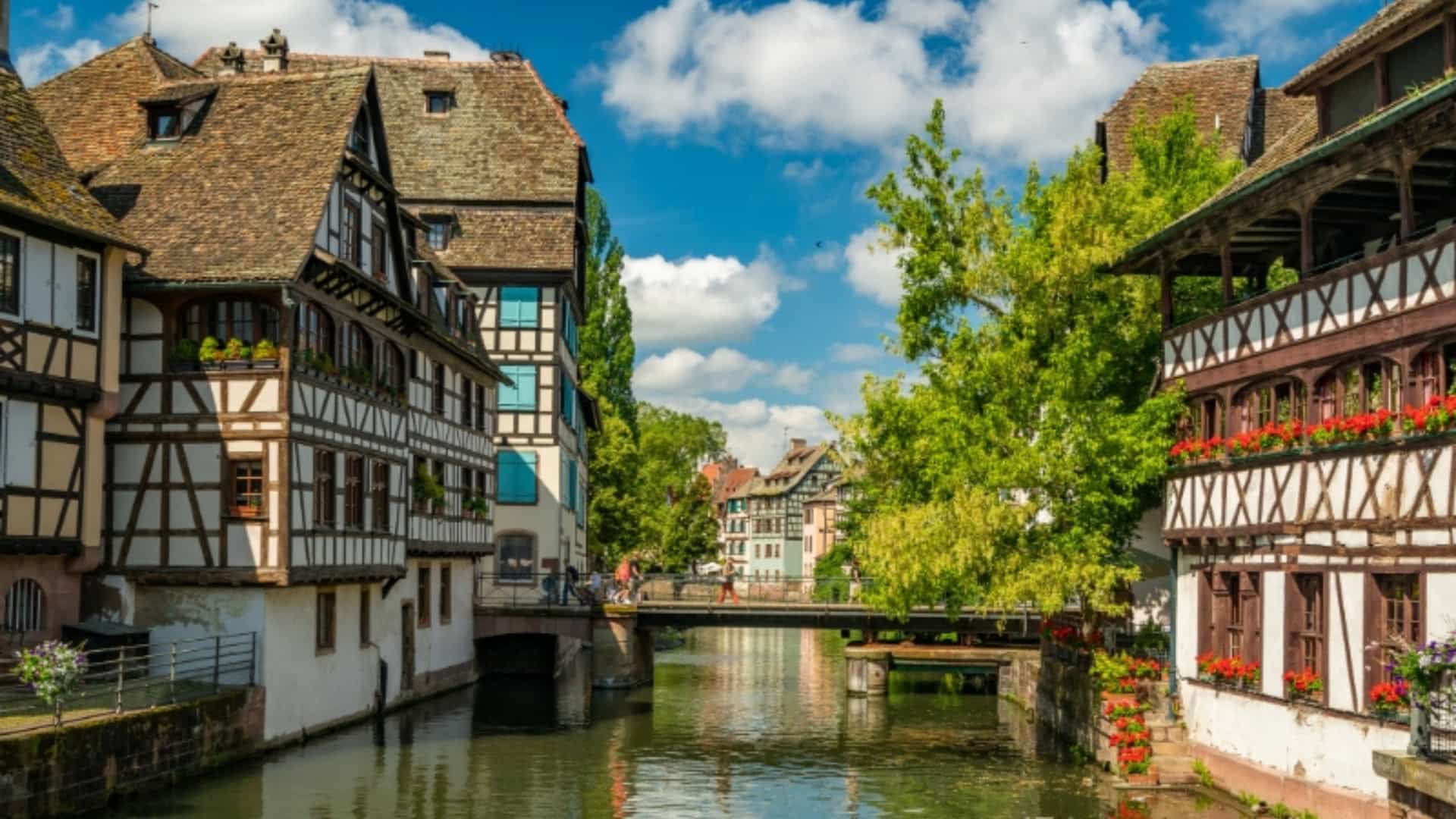 A view of the canal in the Petite France district of Strasbourg, France. The canal is bordered by charming half-timbered houses with dark wood beams and white walls, some decorated with red flowers. A stone bridge crosses the canal, and the sky is blue with fluffy white clouds. The image captures the historic, medieval architecture along a tributary of the Rhine River.