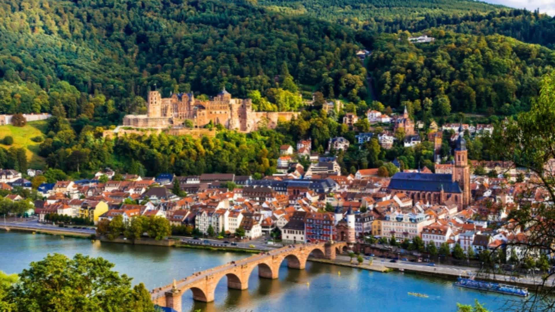 An elevated view of Heidelberg, Germany, showing the old town nestled along the Neckar River, a tributary of the Rhine. The image features the iconic Heidelberg Castle on a forested hill overlooking the city and the Old Bridge spanning the river. The cityscape is filled with historic buildings with red-tiled roofs, and the surrounding hills are covered in lush green trees. The image captures the historic charm and scenic beauty of this Rhine River region.
