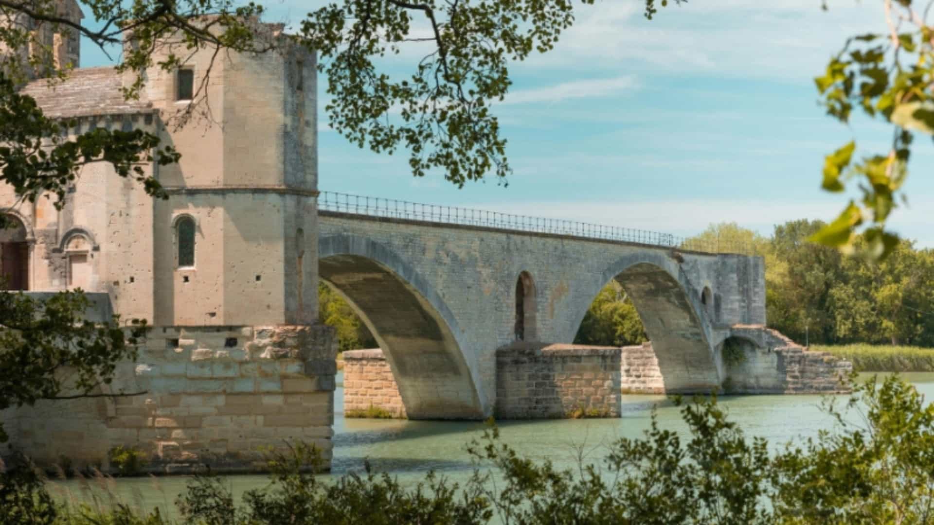 The Pont Saint-Bénézet, a historic stone bridge with a chapel, arches over the Rhone River in Avignon, France. The weathered, light-colored stone of the bridge stands against the green-blue water and a partially cloudy sky. The image captures the historic architecture and scenic views along the Rhone River.