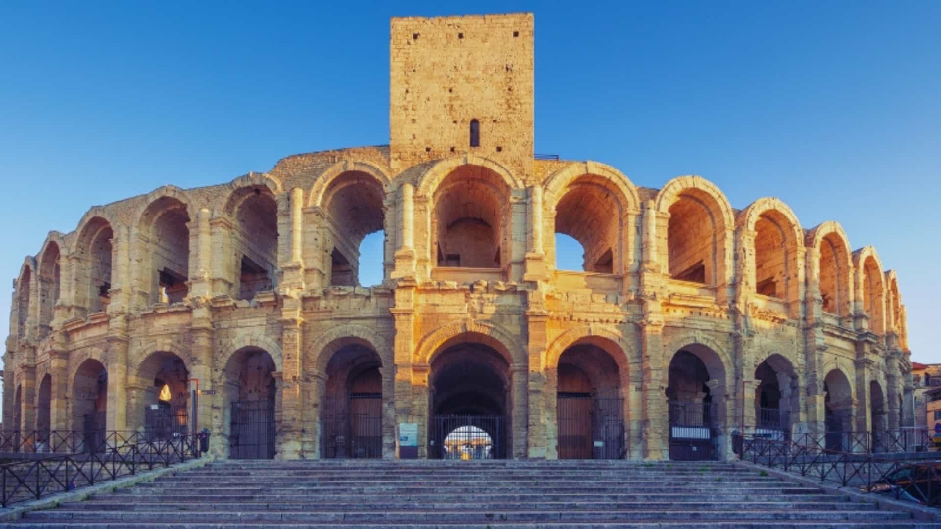 A large, well-preserved Roman amphitheater in Arles, France, with a blue sky in the background. The structure consists of multiple levels of arches and a central watchtower, built from light-colored stone. The image captures the historical significance of the Roman era in the Rhone River region.