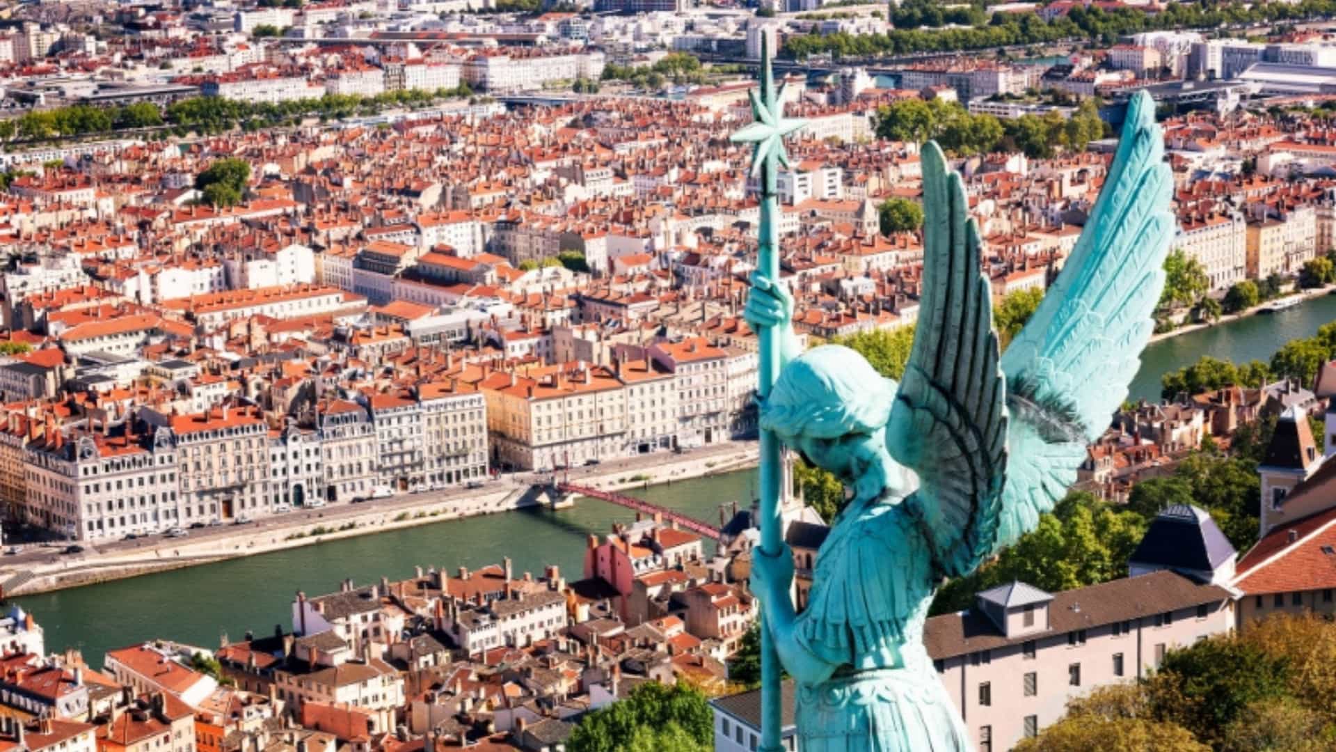 An elevated view of Lyon, France, featuring a large bronze statue of an angel in the foreground. In the background, the Saône River, a tributary of the Rhone, meanders through the cityscape, which is densely packed with buildings with red-tiled roofs. The image captures the urban landscape and art of a major city in the Rhone River region."