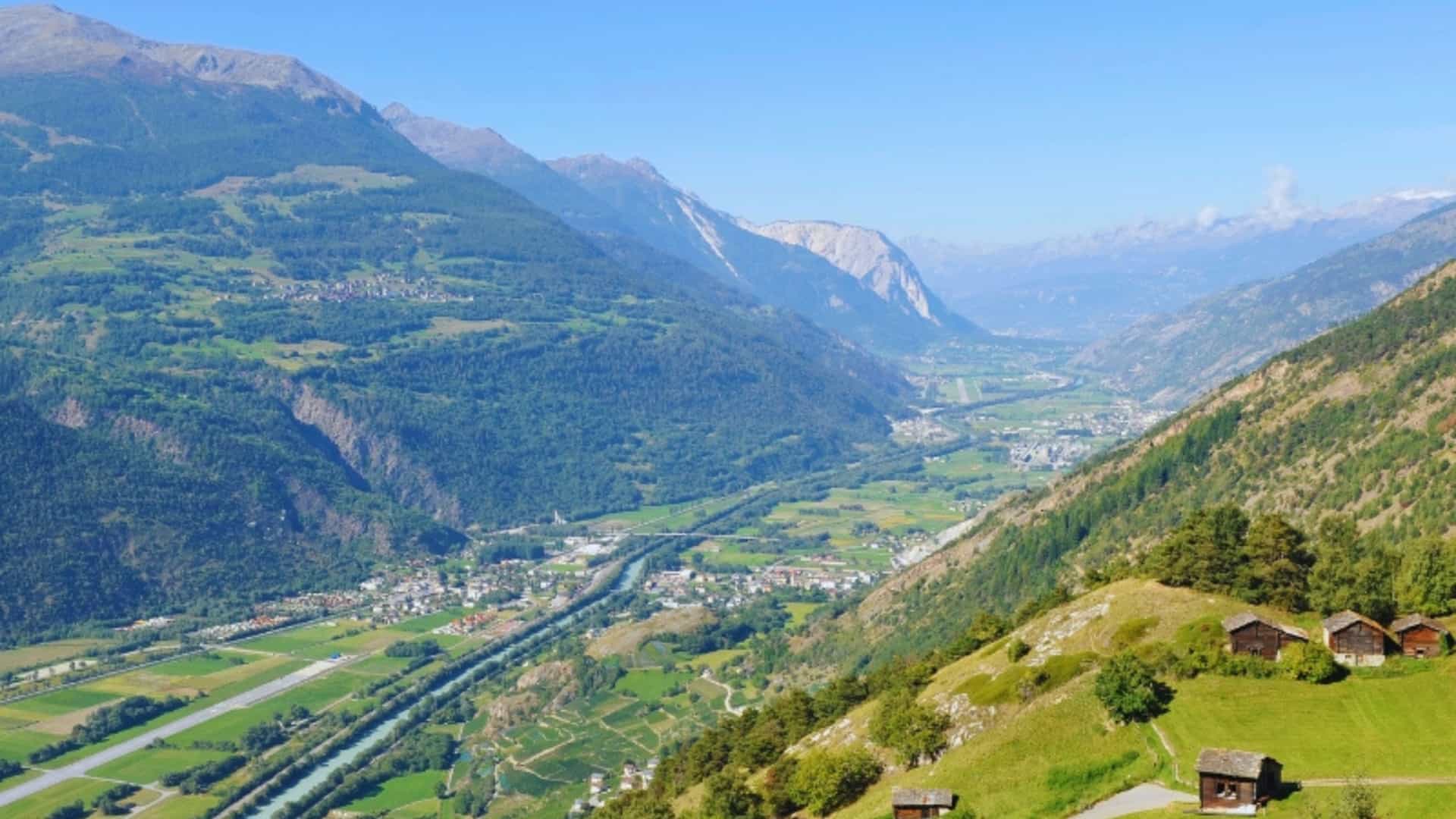 An aerial view of the Rhone River valley in Switzerland, showing the river winding through a wide valley surrounded by towering Alps mountains. Small villages and green fields are visible on the valley floor, while forested hillsides and small wooden chalets dot the slopes. The image captures the natural beauty and scenic landscape of the Swiss portion of the Rhone River.
