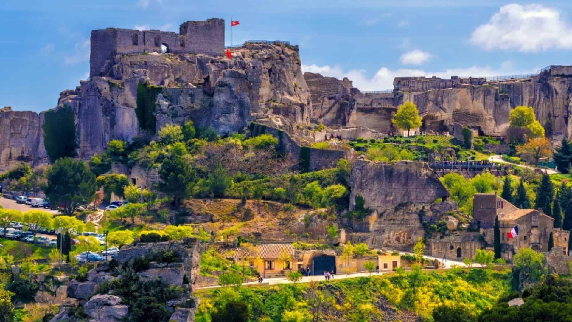 The dramatic ruins of a medieval fortress sit atop a rocky outcrop in Les Baux-de-Provence, France. The stone structure has a flag flying from a tower, overlooking the small village nestled into the hillside below. The landscape is a mix of rugged rock and vibrant green and yellow foliage, all under a bright blue sky. The image captures the historical significance and dramatic landscape of this Rhone River region.