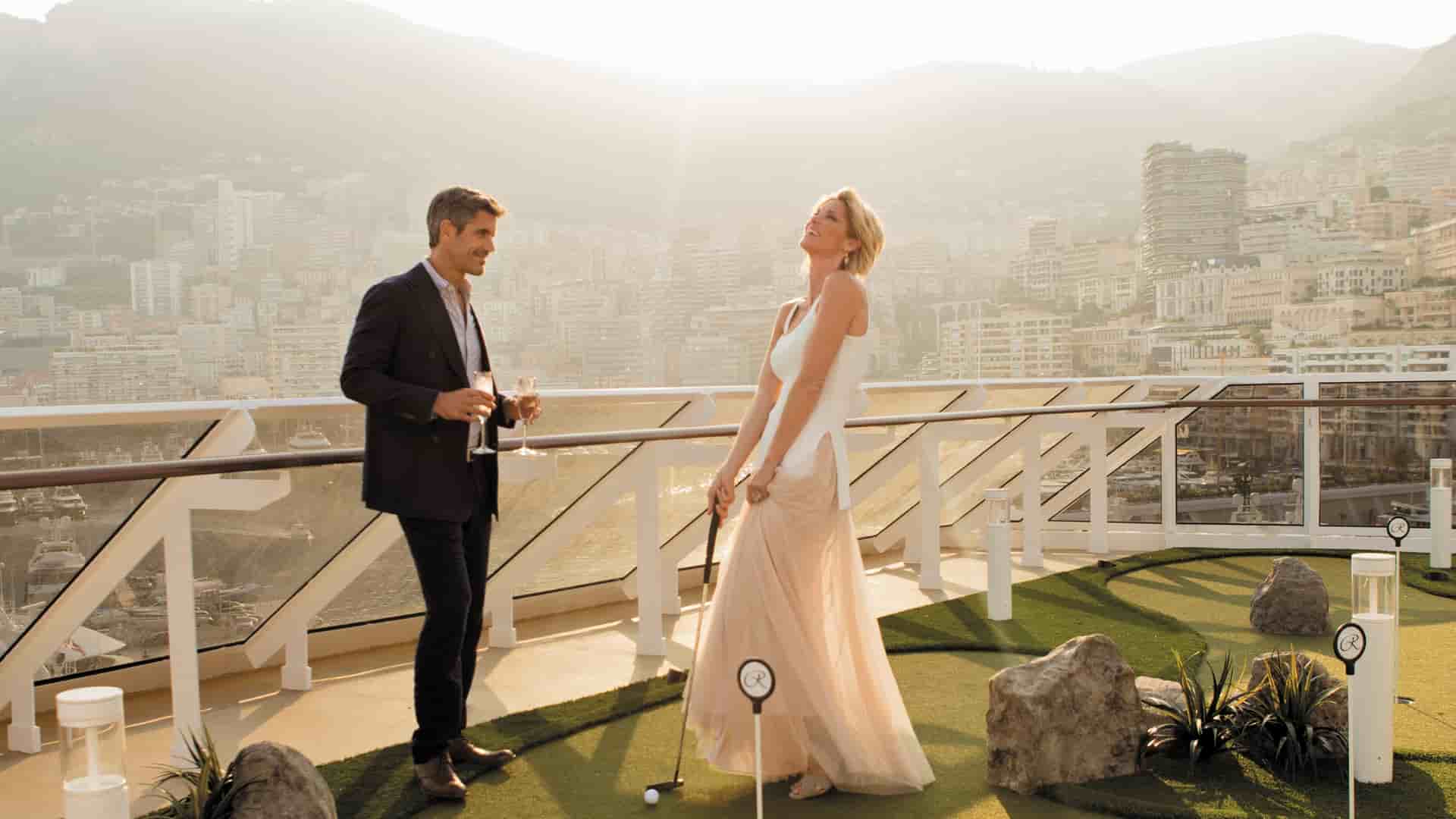 A well-dressed couple laughs while playing mini-golf on the sunlit deck of a luxury Regent Seven Seas cruise ship, with a scenic coastal city nestled into hillsides visible in the background.