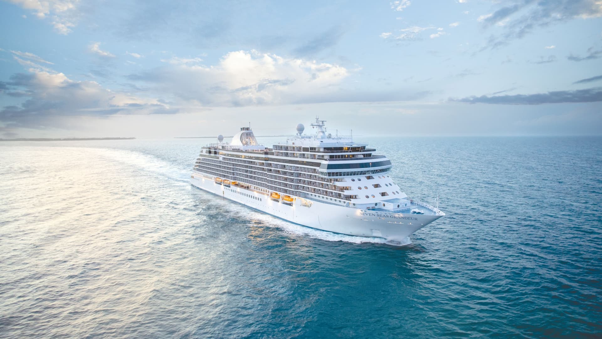 The luxury cruise ship Seven Seas Grandeur sails through the open blue ocean, leaving a white wake beneath a soft, cloudy sky.