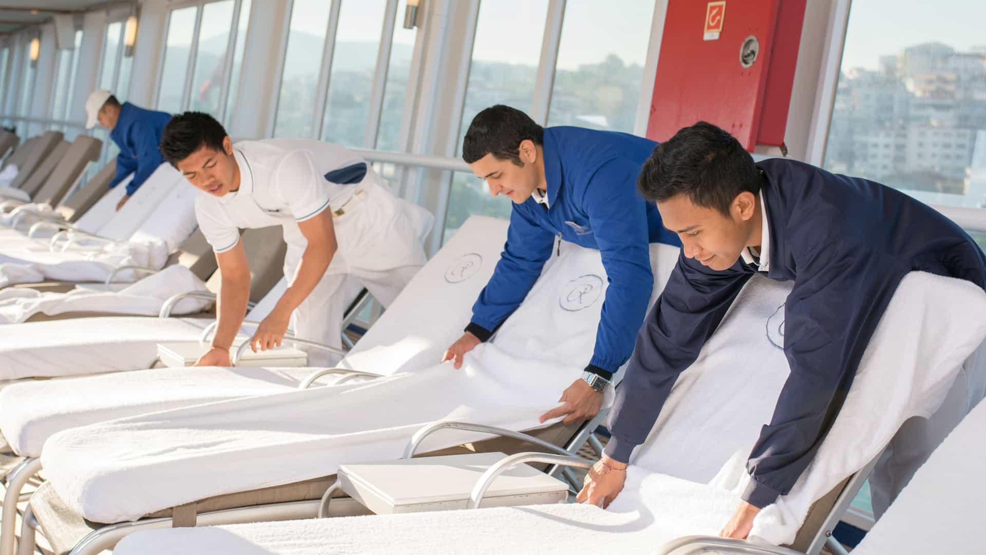 Crew members in uniform carefully arrange white towels on lounge chairs along the sun deck of a cruise ship.
