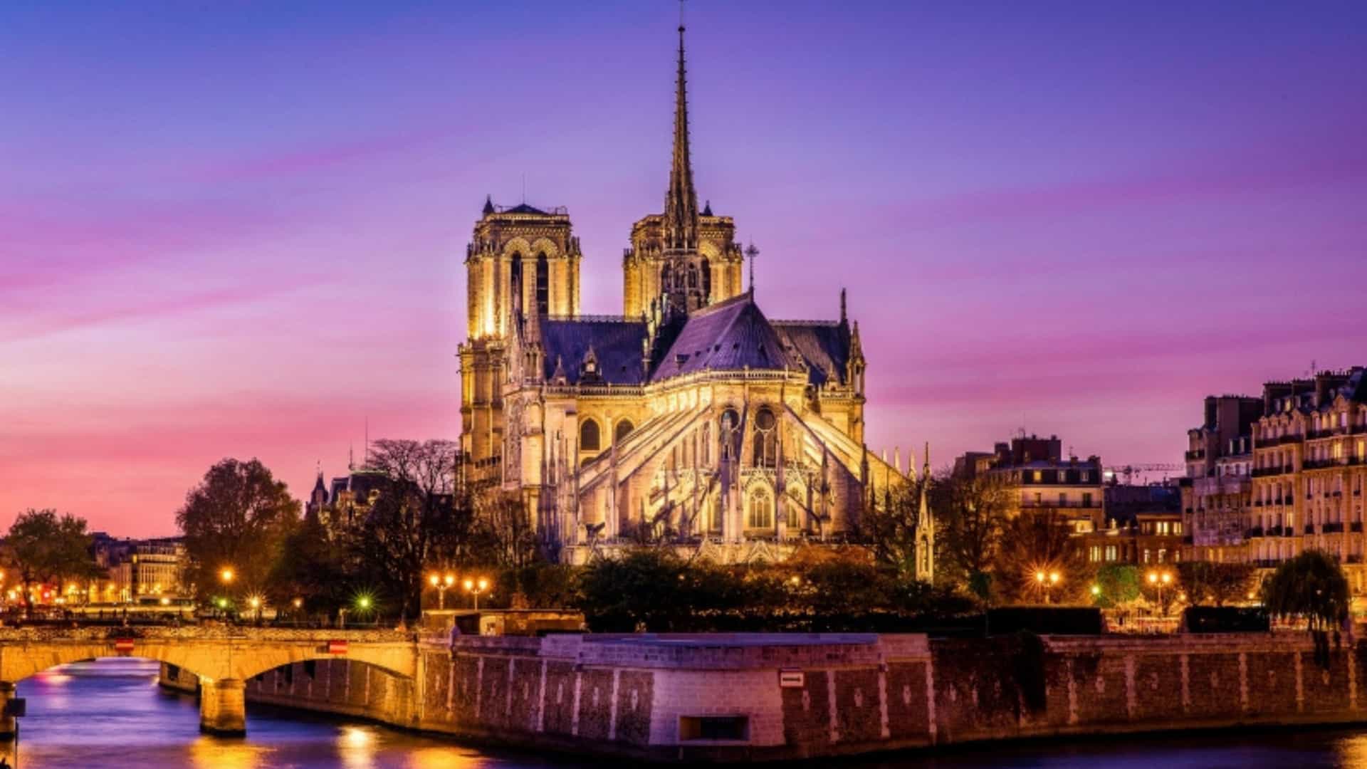The Notre-Dame Cathedral in Paris, France, illuminated at dusk. The iconic Gothic cathedral is positioned at a bend in the Seine River, with a bridge and the city lights reflected on the water's surface. The sky is a dramatic mix of pink and purple, creating a striking scene that captures the essence of this major city's architecture and waterways.