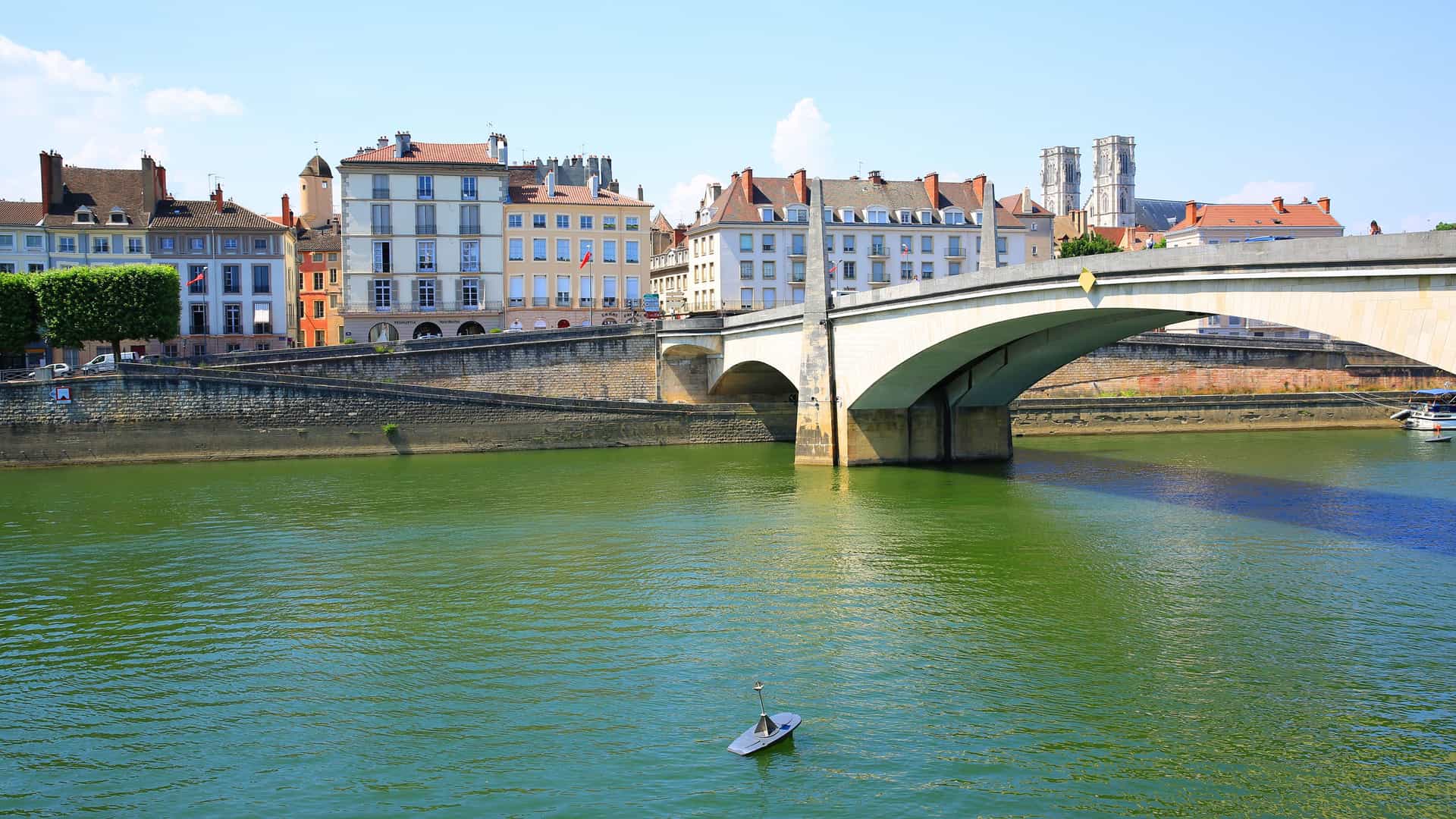 The city of Chalon-sur-Saône, France, is situated on the Saône River, with a large, arched stone bridge connecting two sides of the city. The riverbank is lined with historic buildings with colorful facades. The water is a murky green, and a small, lone paddleboard with a paddle lies in the middle of the river. The image captures the urban landscape along the Saône River.