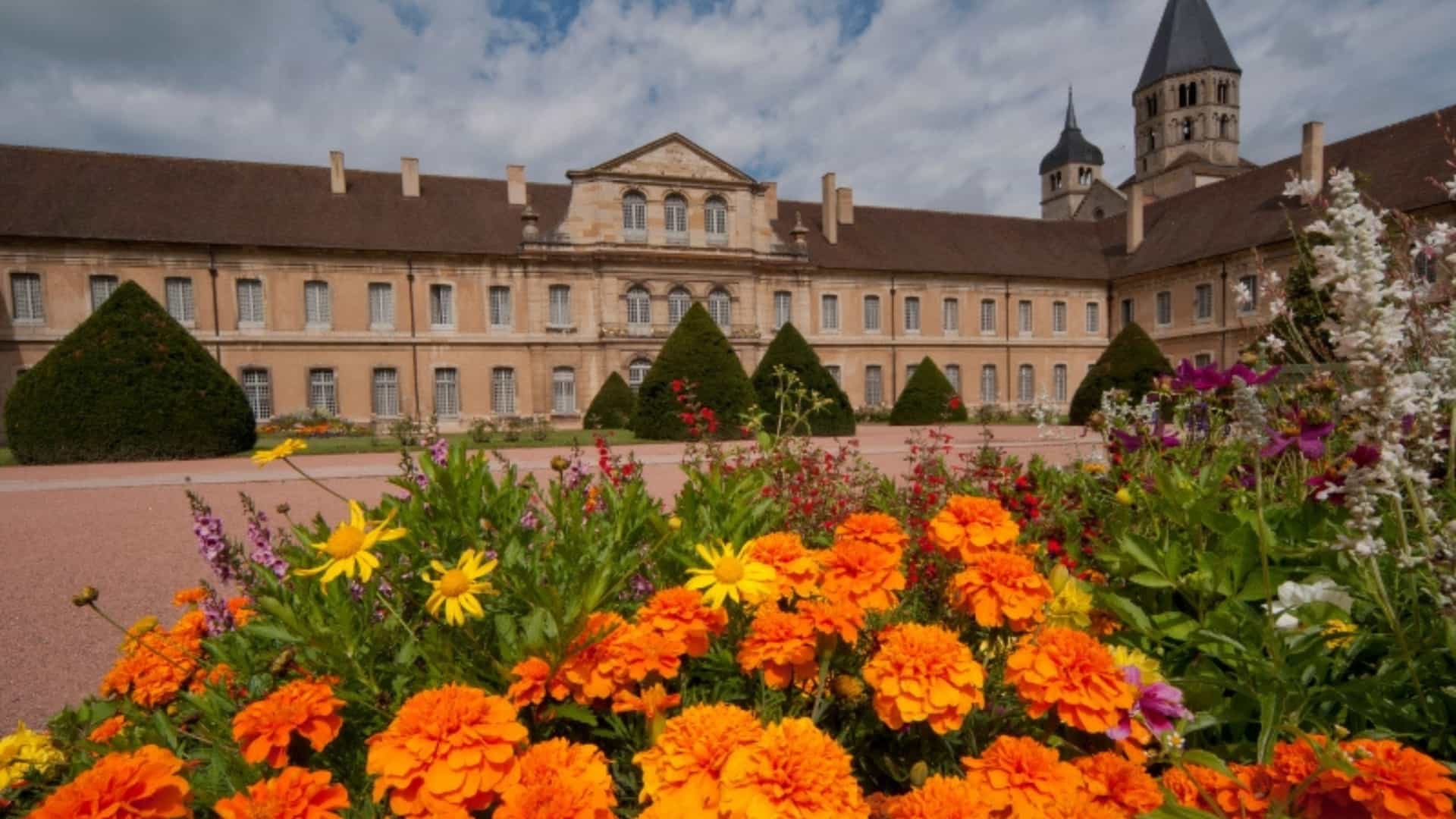 The historic Cluny Abbey in France, a famous monastery and garden near the Saône River. The image shows a manicured flowerbed with bright orange marigolds and other colorful flowers in the foreground. In the background, the large, ornate stone building of the abbey and its steeple stand under a partly cloudy sky, showcasing the historic and religious significance of this region.