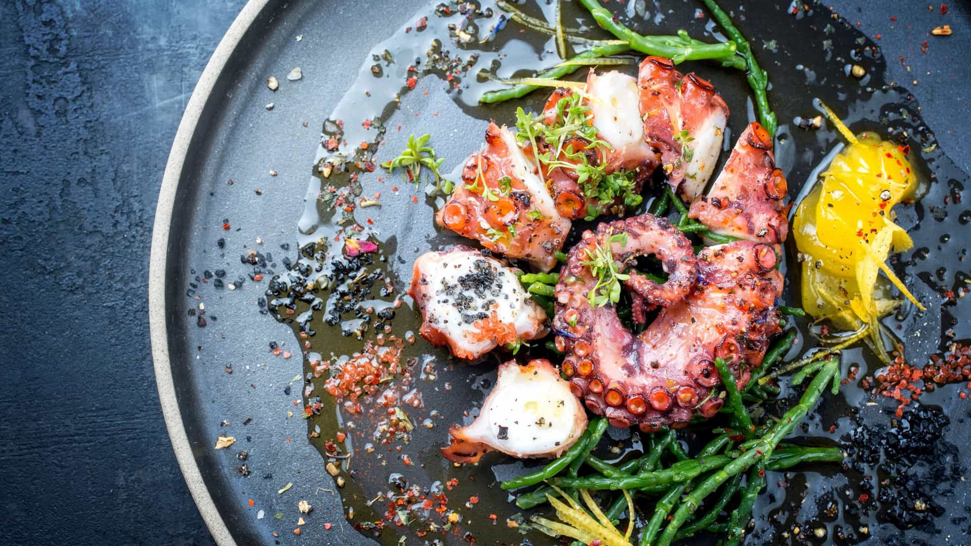 Overhead view of sliced and whole grilled octopus tentacles on a dark plate with green sea vegetables, microgreens, a citrus garnish, black salt and red spice.