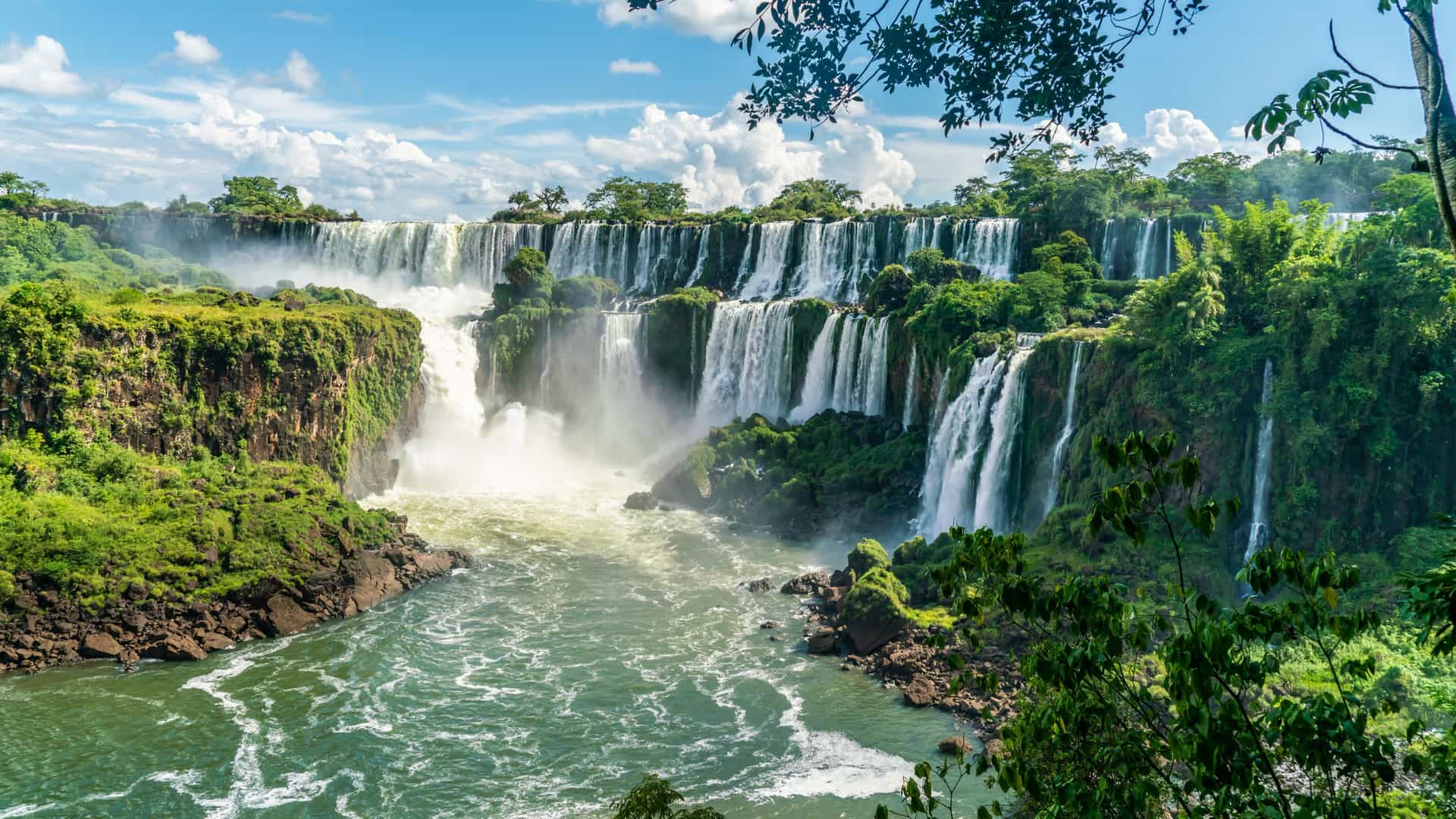 A panoramic view of the multi-tiered Iguazu Falls in Argentina, featuring massive white waterfalls cascading over lush green cliffs into a swirling river below, framed by tropical foliage under a bright blue sky.