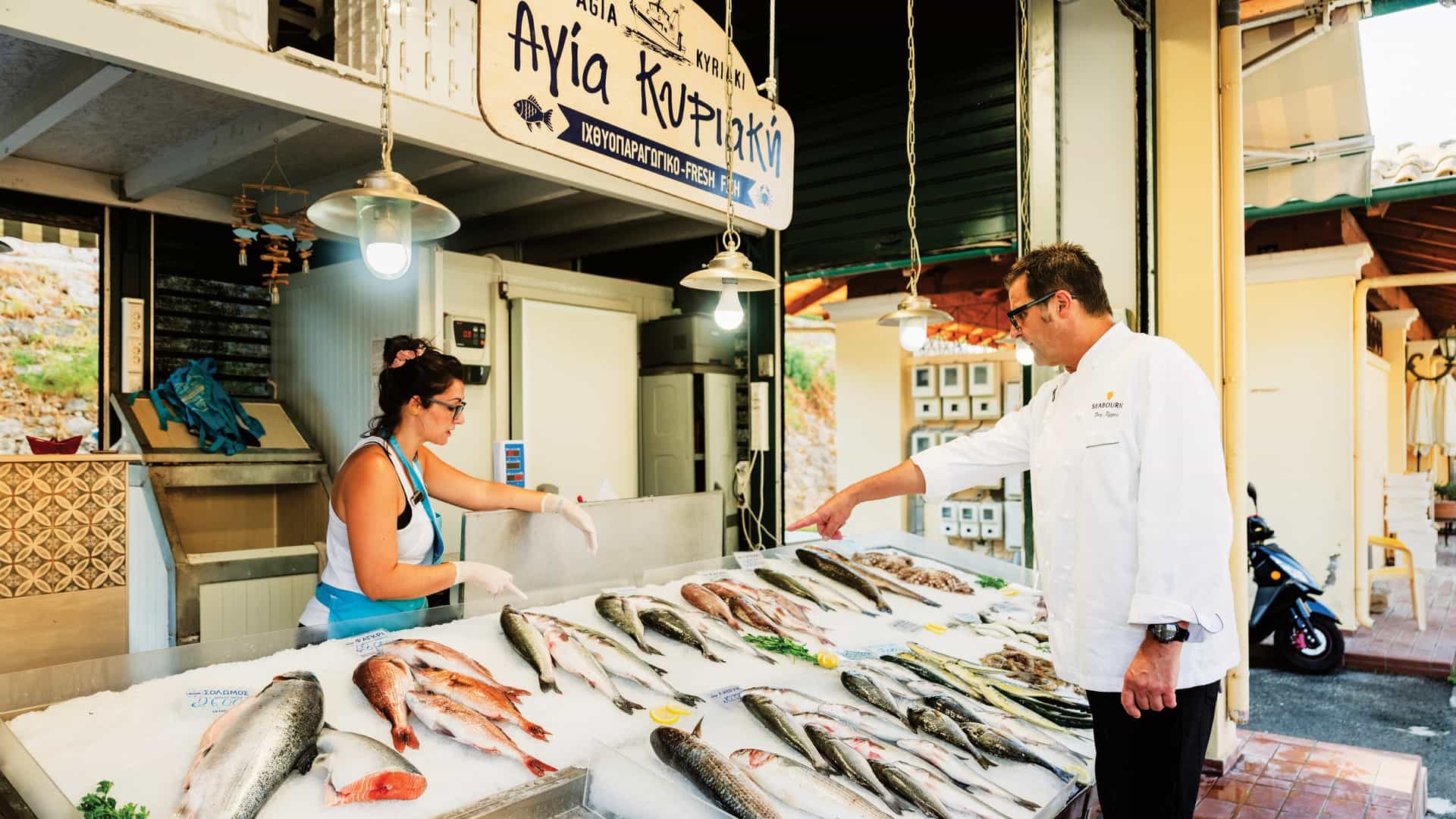 Seabourn Shopping with the Chef excursion in Greece. A luxury cruise chef selecting fresh, local seafood at a traditional Mediterranean fish market for onboard dining.