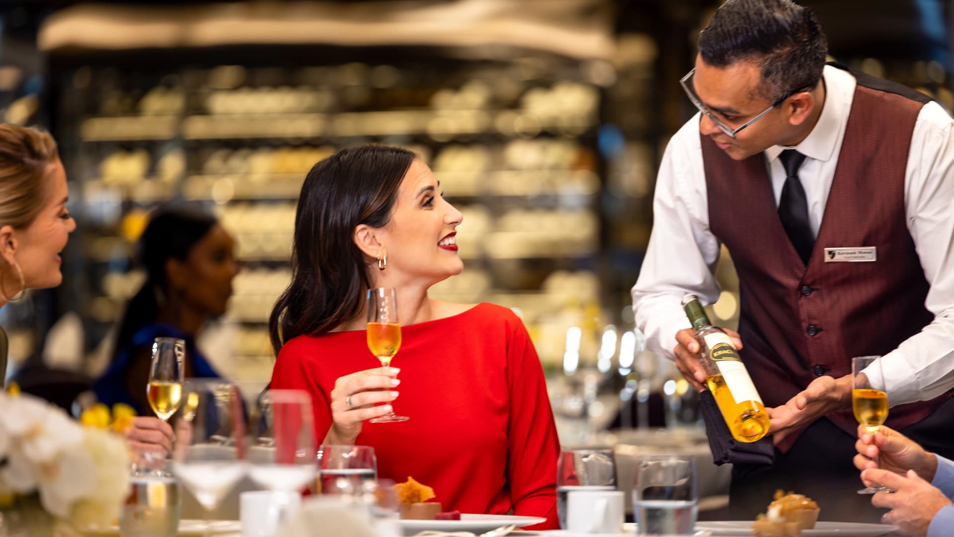 A woman in a red long sleeved dress, aboard Seabourn Venture, sits at a dining table with a glass of wine held in her right hand, smiling at the waiter who is displaying a glass of wine for her to see.