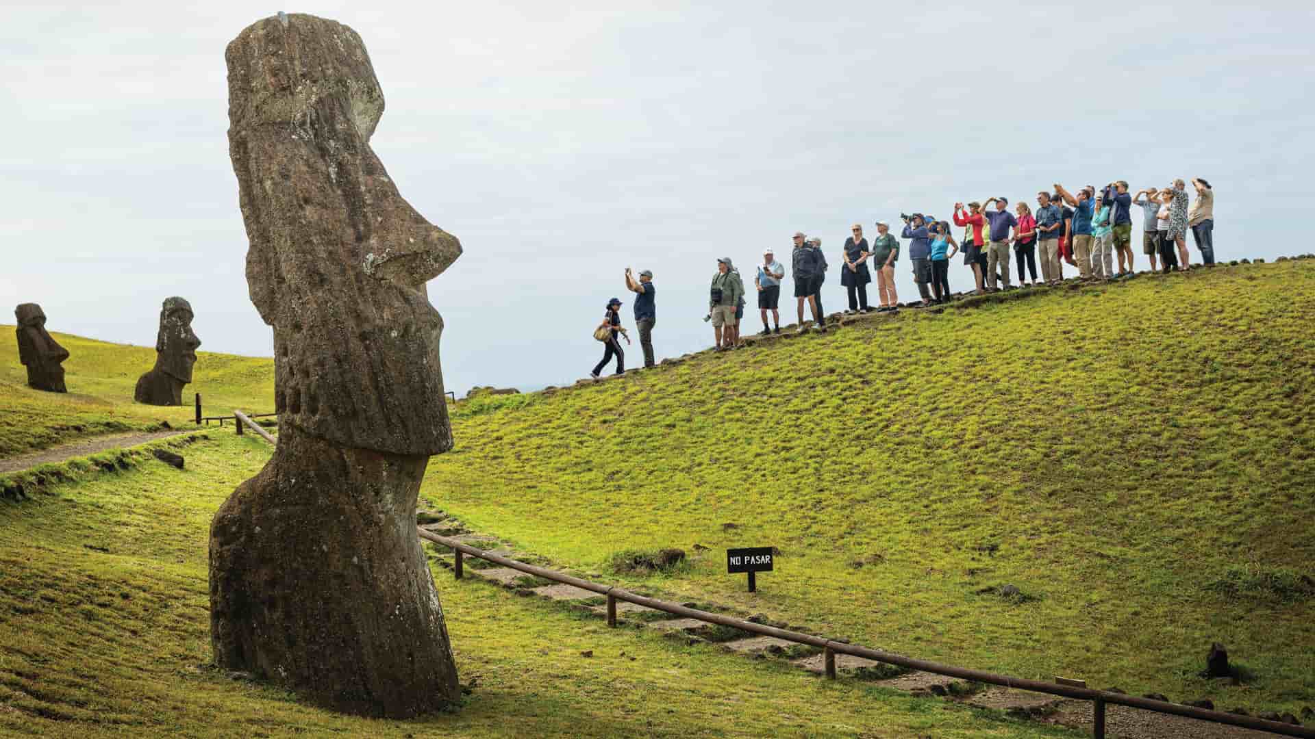 Alt Text Tourists standing along a grassy slope on Easter Island, Chile, viewing monolithic Moai stone statues under an overcast sky.