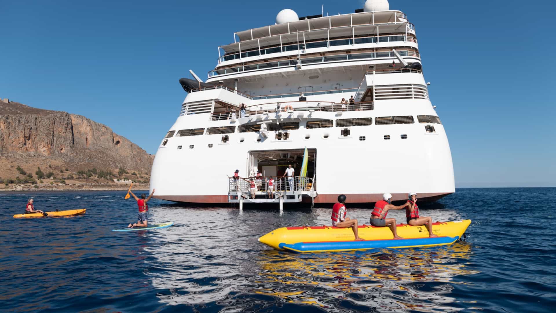 The stern of Seabourn Cruise Line's Seabourn Ovation ship is docked at sea, with a group of people enjoying watersports from the ship's marina, including a man on a stand-up paddleboard and two groups on banana boats.