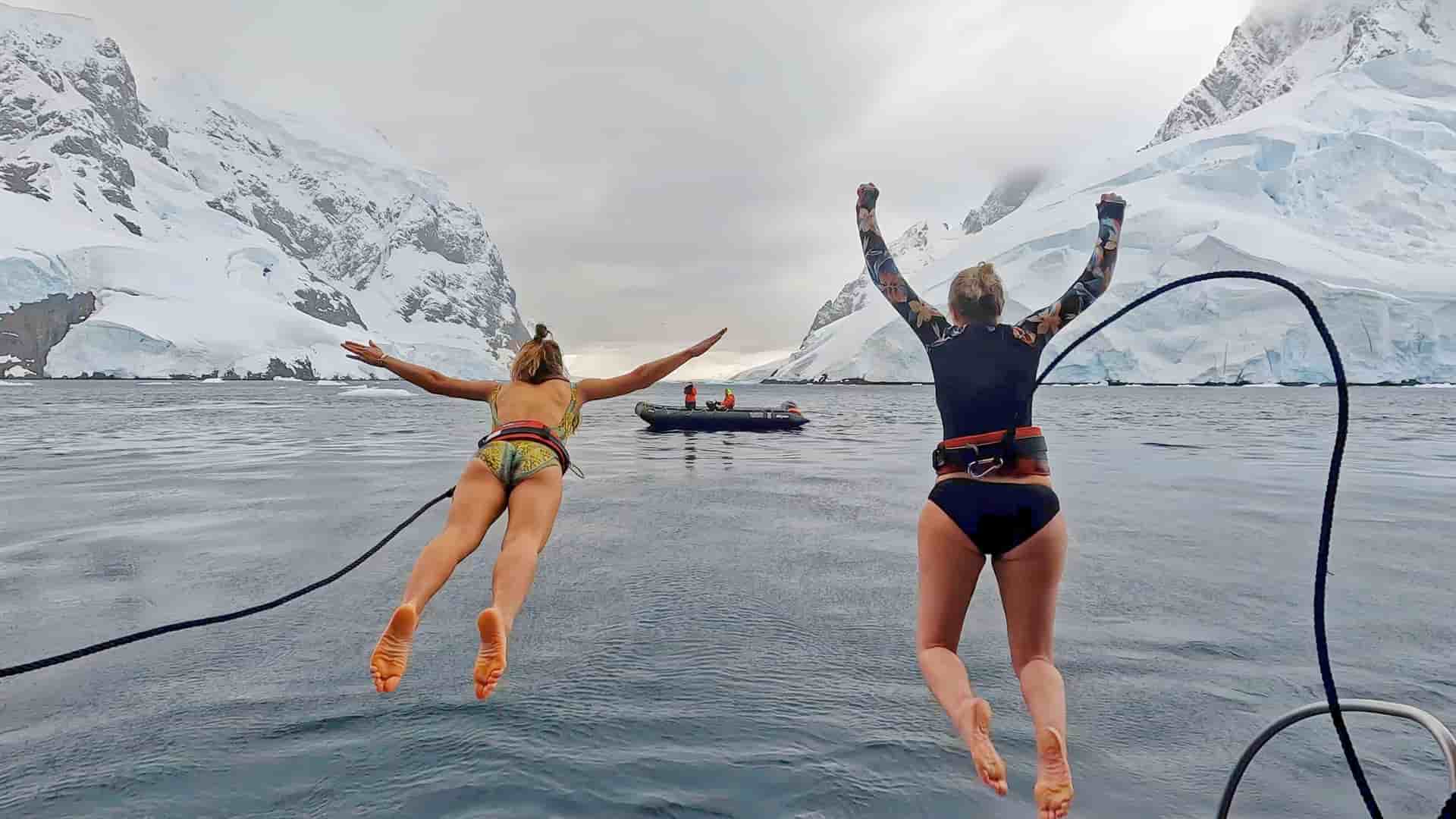 Two people in swimsuits and harnesses jump into the water from the Seabourn Cruise Line's Seabourn Venture ship, with snow-covered mountains in the background.