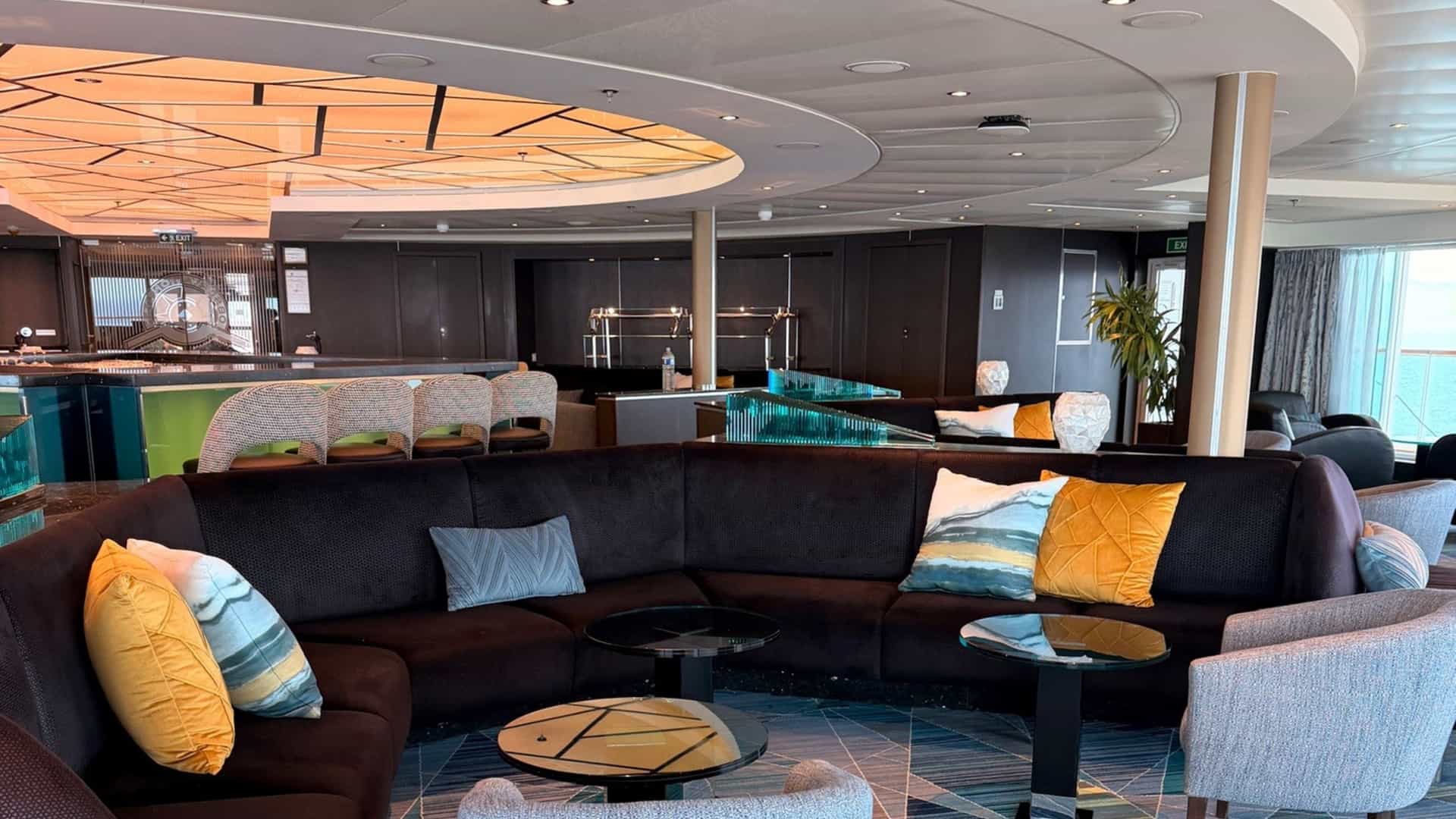 The refreshed Observation Bar onboard the Seabourn Quest, featuring a dark curved sectional sofa with yellow and blue accent pillows, small round coffee tables, and bar seating in the background.