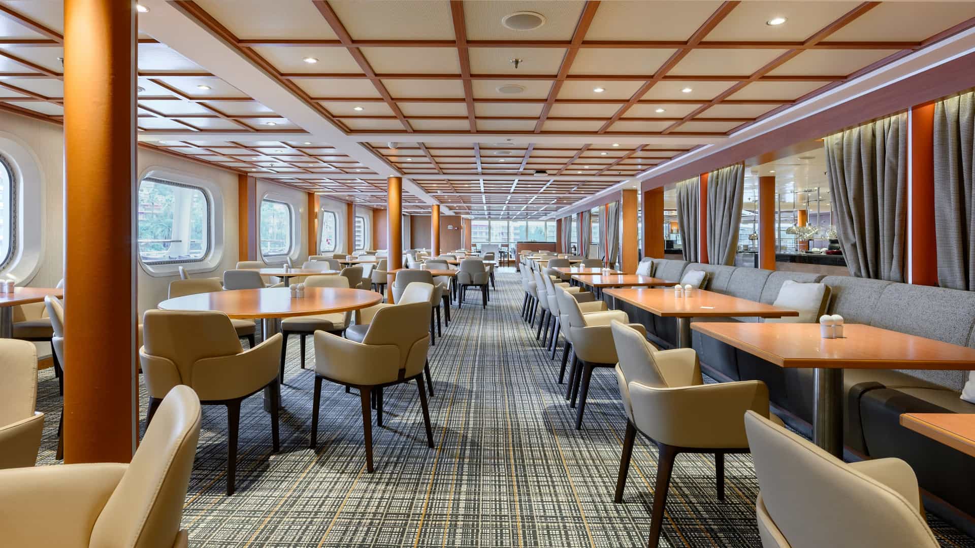The Colonnade restaurant onboard the Seabourn Quest after it's renovation, featuring wooden dining tables, light beige armchairs, banquette seating, and a decorative paneled ceiling.