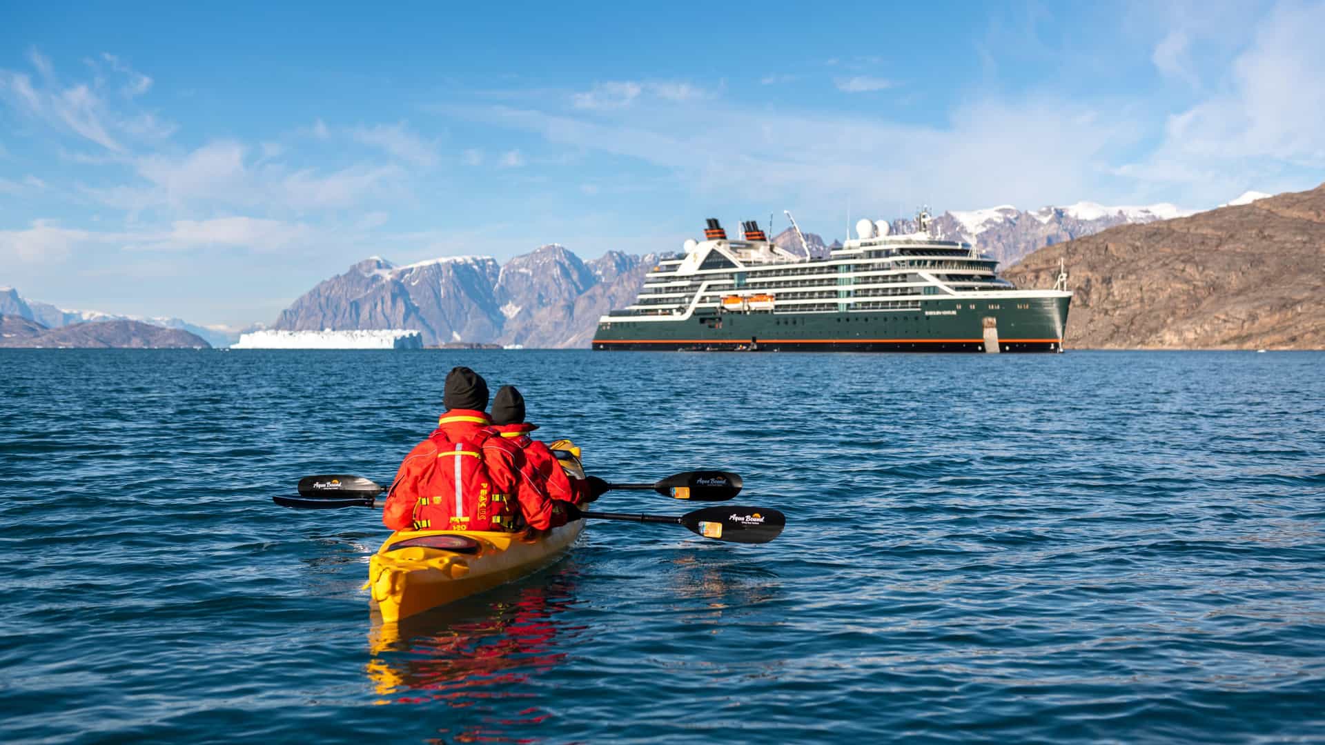 Two people kayak in the waters of Greenland with a Seabourn Venture cruise ship and snow-capped mountains in the background.