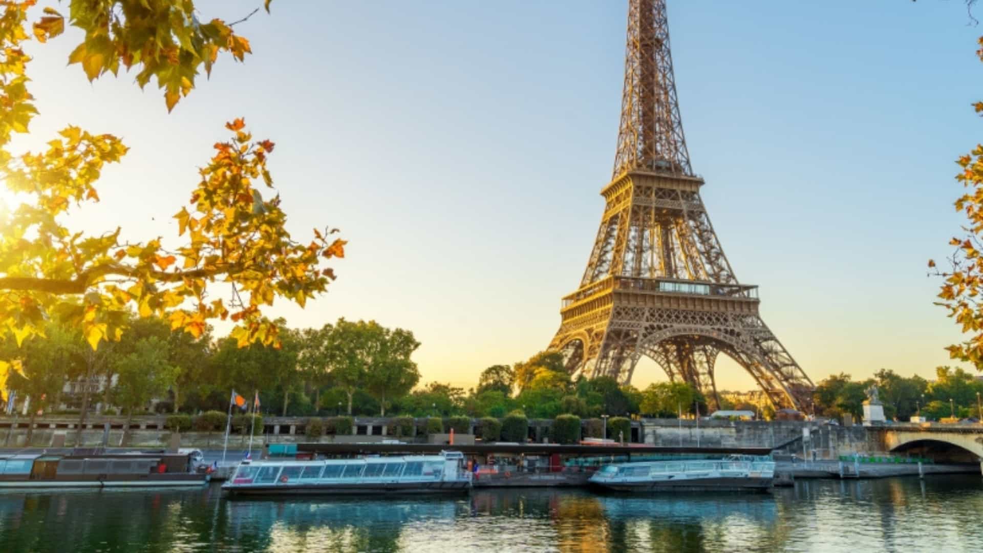 A beautiful view of the Eiffel Tower in Paris, France, from the banks of the Seine River. The sun sets behind the tower, casting a golden glow on the scene and highlighting the autumn leaves of a tree in the foreground. Several tour boats are docked along the river, their reflections visible on the water's calm surface. The image captures a classic and romantic Parisian scene at sunset.