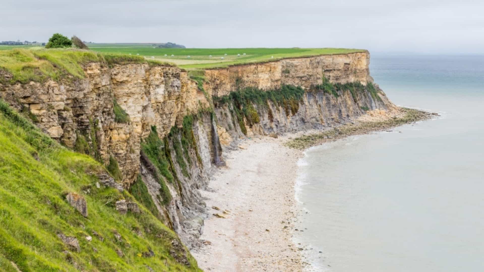 A dramatic view of the high, rugged cliffs of Pointe du Hoc on the Normandy coast of France. The image shows the steep, rocky cliffs dropping down to a narrow pebble beach below, with the calm, gray-blue water of the English Channel visible at the edge. The top of the cliffs is covered in green grass, with a field in the distance. The scene is a powerful reminder of its historical significance from the D-Day landings during World War II.