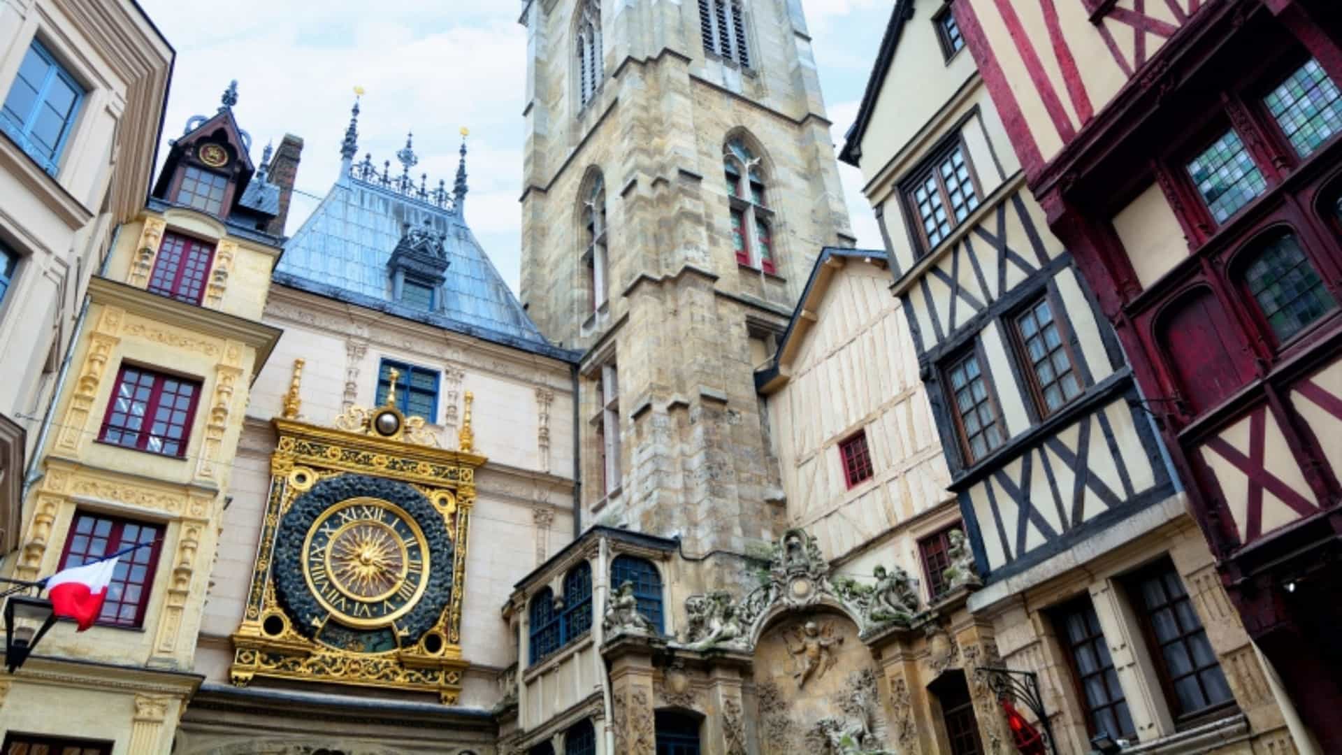 A low-angle shot of the Gros Horloge, a famous astronomical clock, in Rouen, France. The ornate, golden clock face is mounted in a Renaissance archway that connects two half-timbered buildings with intricate woodwork and traditional windows. Above the clock, a Gothic bell tower rises, with a spire at its peak. The image captures the historic architecture and charm of the city of Rouen, located along the Seine River.