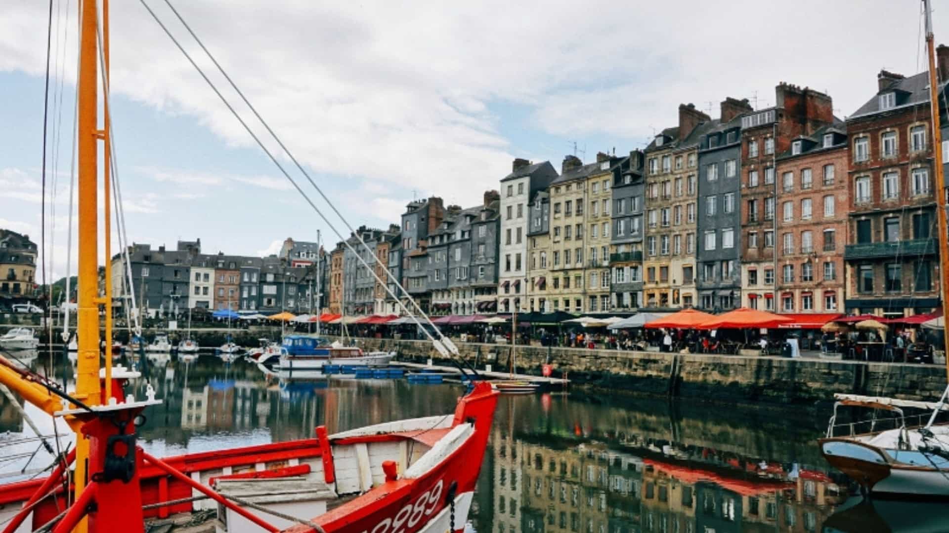 A picturesque view of the old harbor in Honfleur, France, on a cloudy day. The harbor is filled with small boats, including a prominent red fishing boat in the foreground. The water is calm and reflective, mirroring the tall, narrow, and colorful buildings that line the quayside. The buildings feature traditional Norman architecture with half-timbered facades and slate roofs. In the background, people are visible sitting at tables outside of cafes and restaurants, capturing a lively scene in this charming Normandy port town.