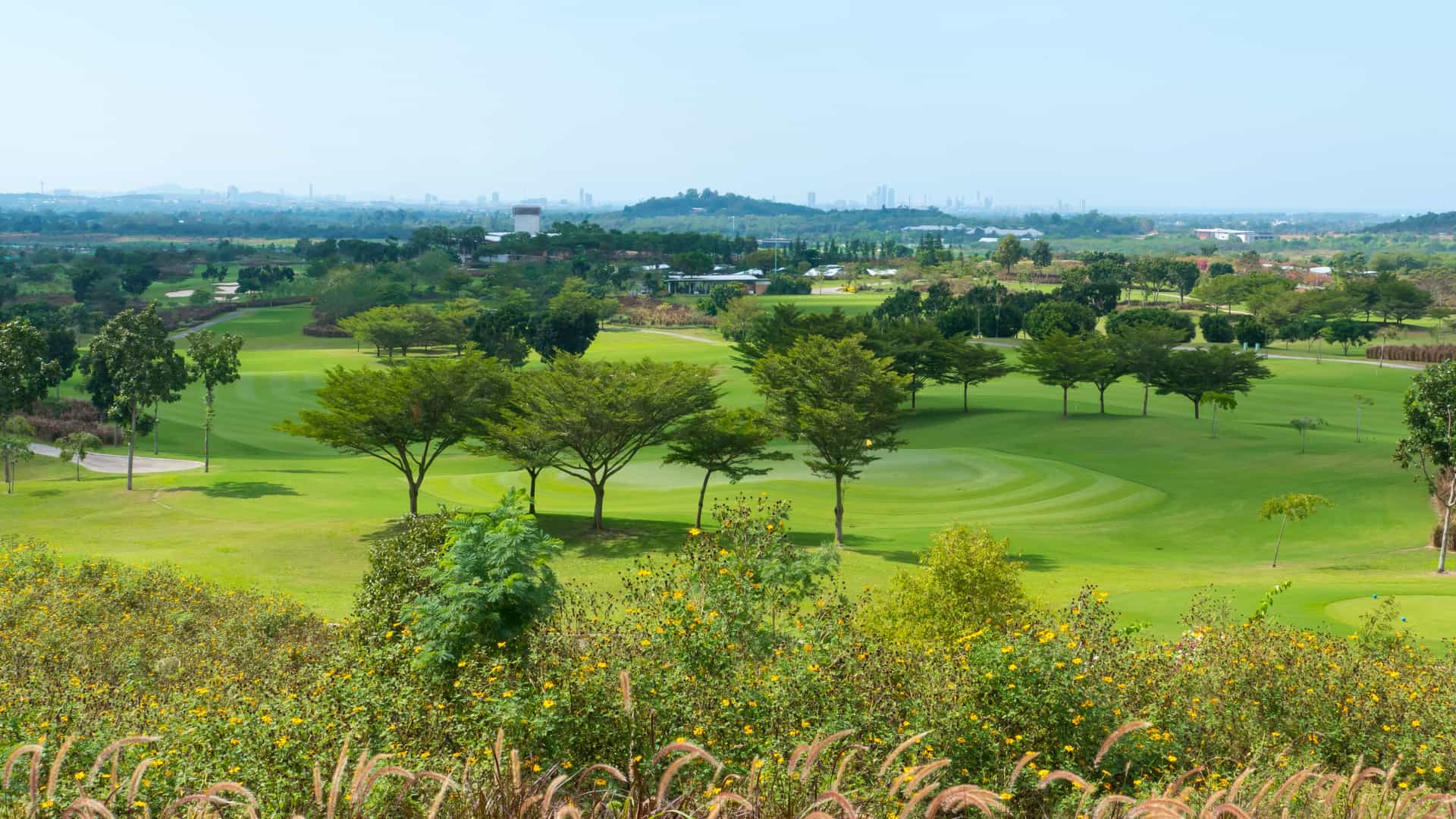 A wide panoramic view of Siam Country Club's Plantation Course in Pattaya, Thailand, featuring rolling green fairways, tropical trees, and a distant city skyline view.