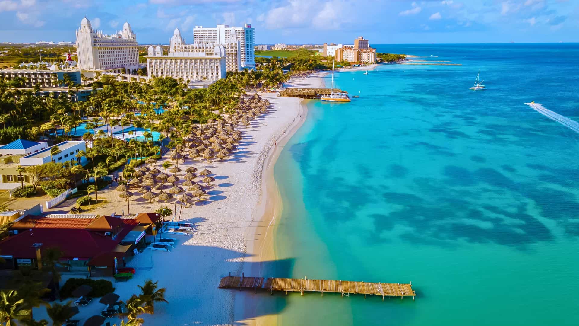 An aerial view of Palm Beach in Aruba, featuring a white-sand beach, resort hotels, and a pier on the turquoise waters of the Southern Caribbean, a destination for Silversea cruises.