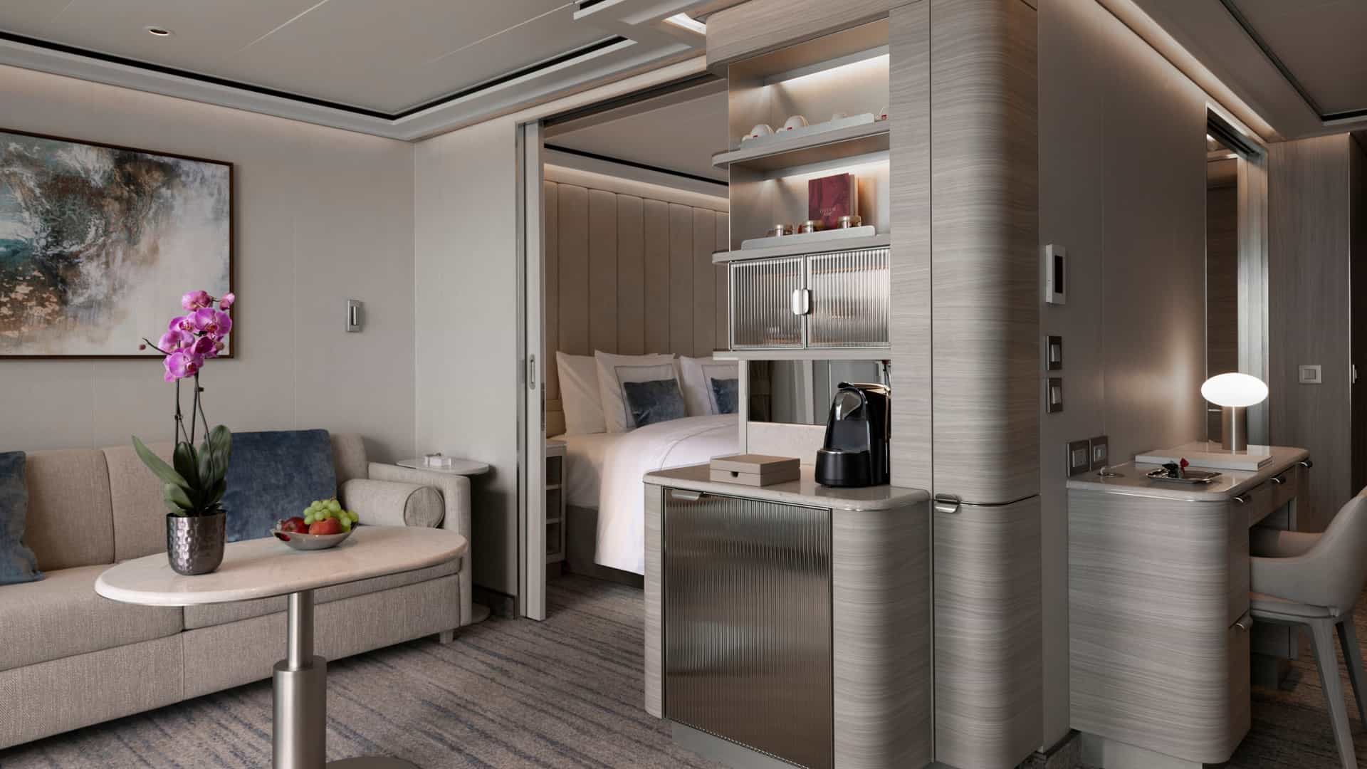 An elegant suite interior with a sitting area containing a sofa and a coffee table adorned with flowers. A sliding glass partition opens into the bedroom area, while a built-in cabinetry unit holds a coffee maker and refreshment station.