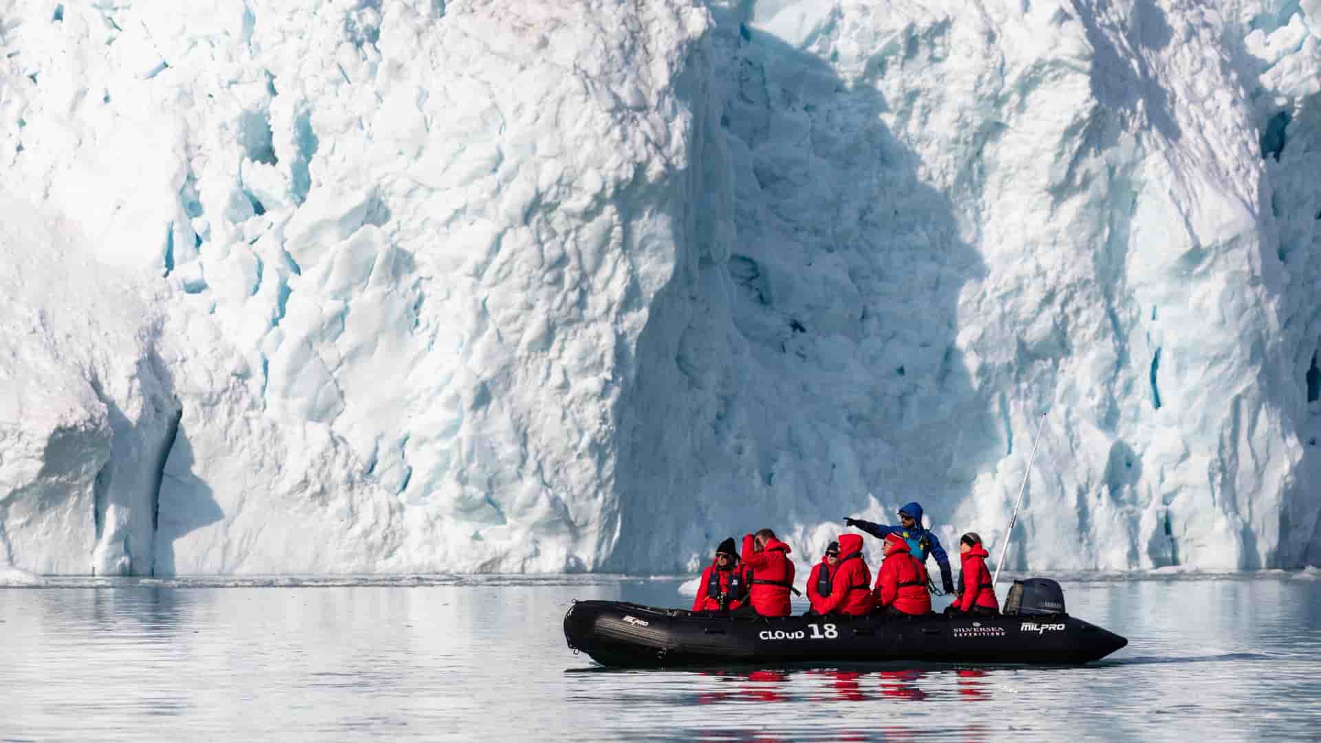 A small Zodiac boat carrying passengers in red parkas navigates dark, choppy waters beneath a towering, rugged stone sea arch in Svalbard.