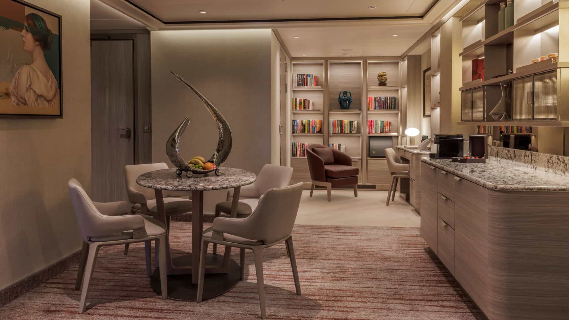 A sophisticated living and dining space featuring a round marble table surrounded by four armchairs. A sculptural metal centerpiece rests on the table, and a long refreshment counter with a built-in coffee bar runs along the right side.