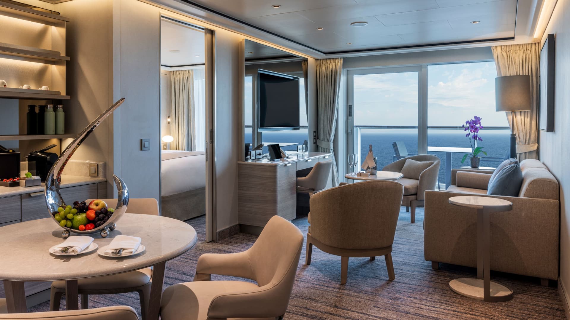 A luxurious cruise suite living area featuring a round dining table with a sculptural fruit bowl in the foreground. To the right, a comfortable seating area sits beside large windows overlooking the ocean, while a sliding door opens to a serene bedroom in the background.