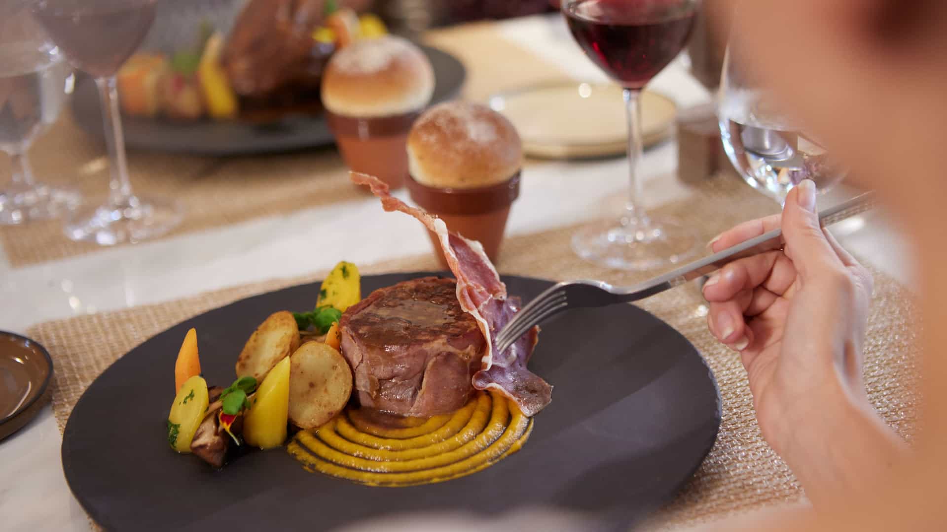 A close up shot over the shoulder of a woman dining at the S.A.L.T. Kitchen aboard Silversea, eating a contemporary steak dish with potatoes.