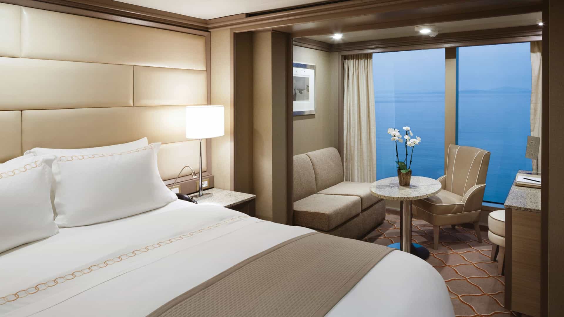 A close-up view of a comfortable bed with crisp white linens and a padded headboard. In the background, a cozy seating area features a sofa, a small round table, and a large window looking out onto the sea.