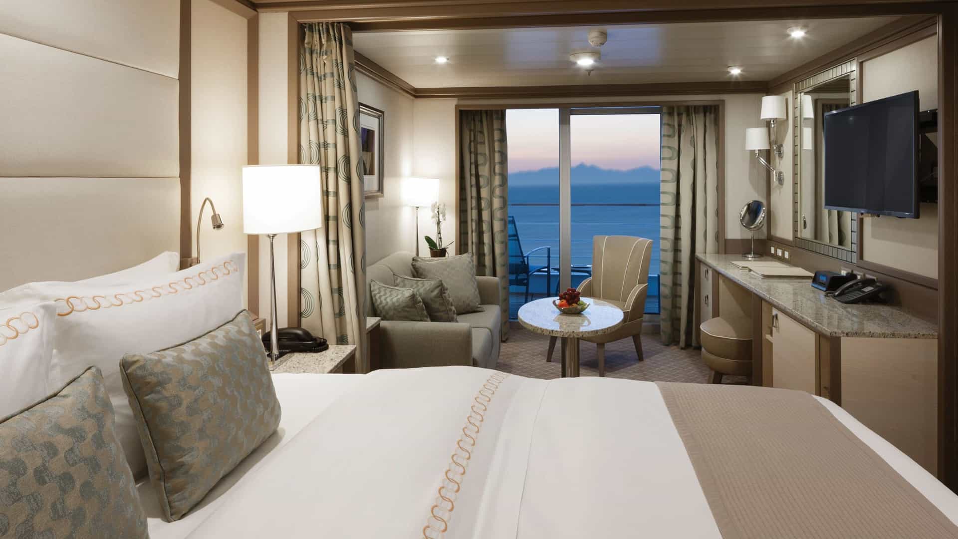 A luxury cruise suite bedroom focusing on a neatly made bed in the foreground. Beyond the bed, a living space includes a sofa, a coffee table, a wall-mounted television, and a sliding glass door opening to an ocean-view veranda at dusk.