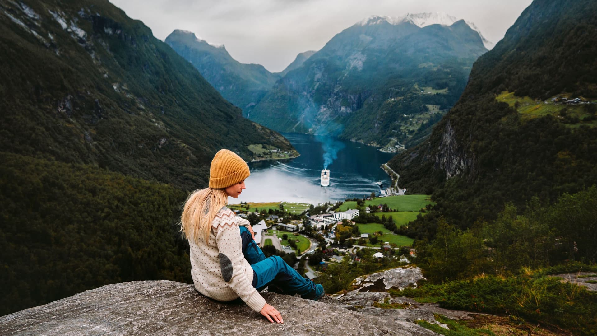 A luxury cruise ship dwarfed by the massive green cliffs of Geirangerfjord, Norway, as seen from the iconic Flydalsjuvet viewpoint.